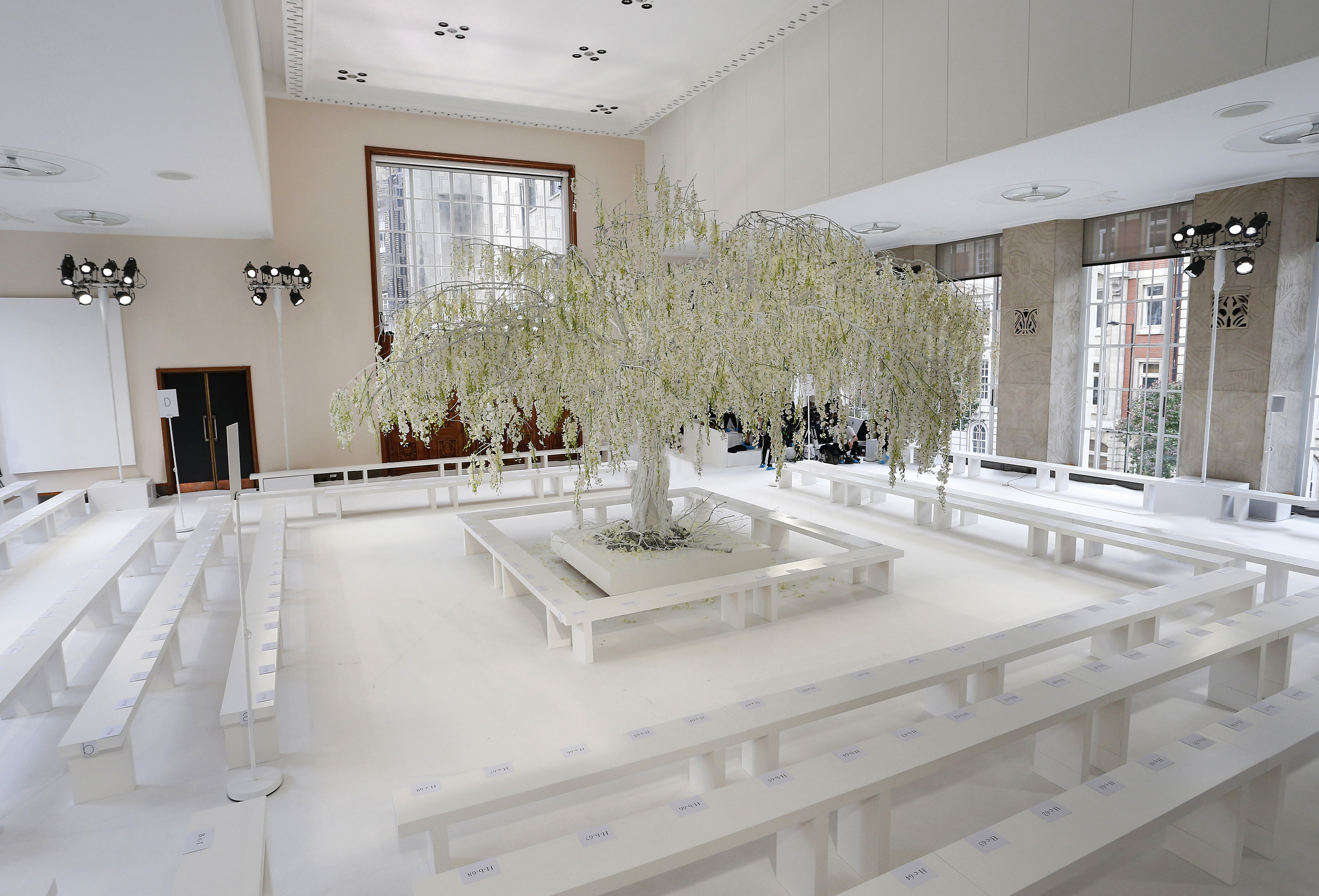 Sala blanca con un sauce de glicinias blancas en el centro de la pasarela y bancos para el público, desfile Delpozo Londres SS 2019 producido por ESMA Events