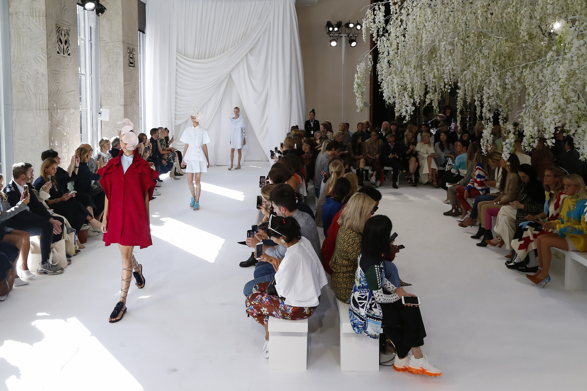Modelo con abrigo rojo avanzando por la pasarela blanca entre el público, desfile Delpozo Londres SS 2019 producido por ESMA Events