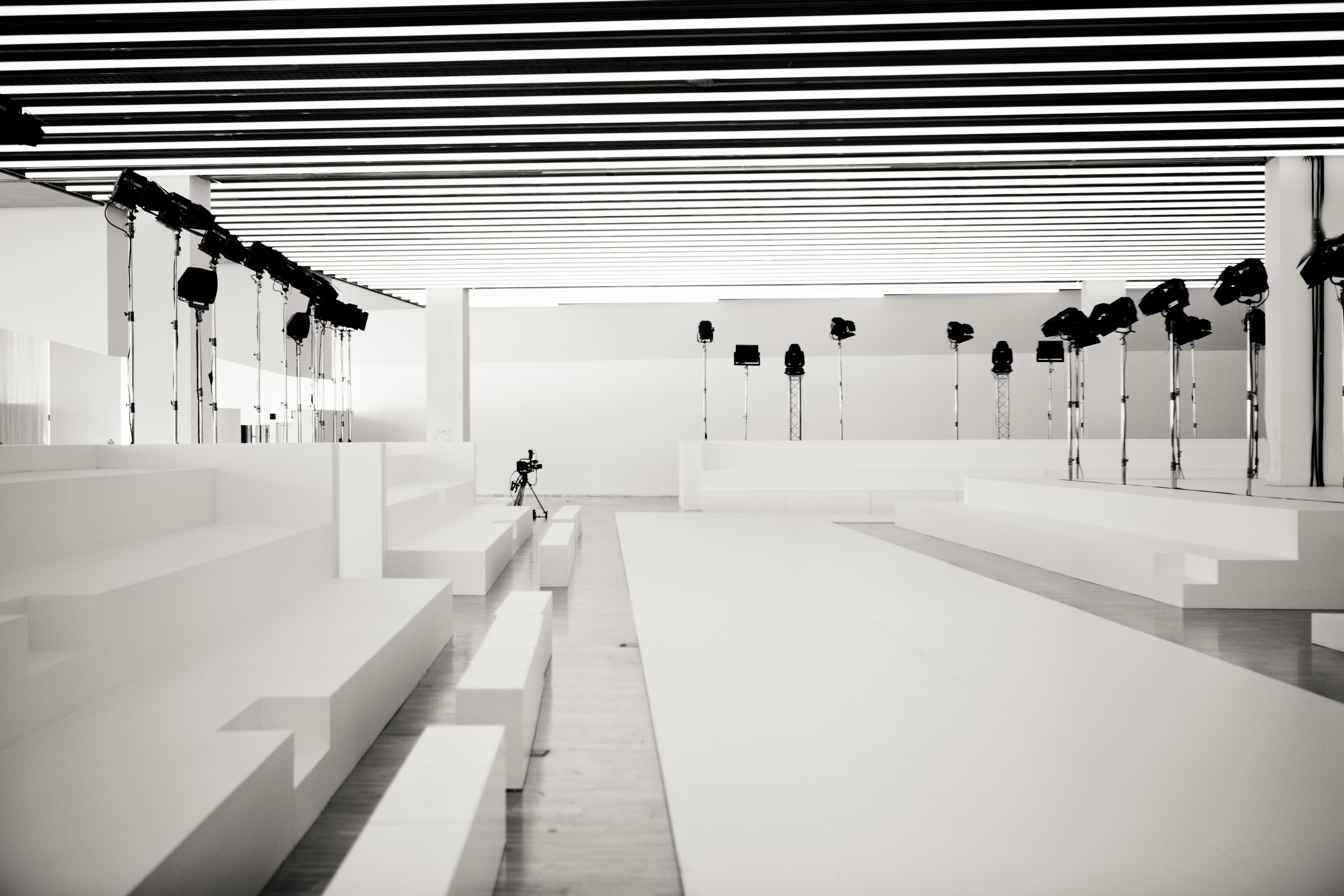 Pasarela blanca flanqueada por gradas blancas y focos sobre trípodes, en blanco y negro, 080 Barcelona Fashion SS 2014 producida por ESMA Events