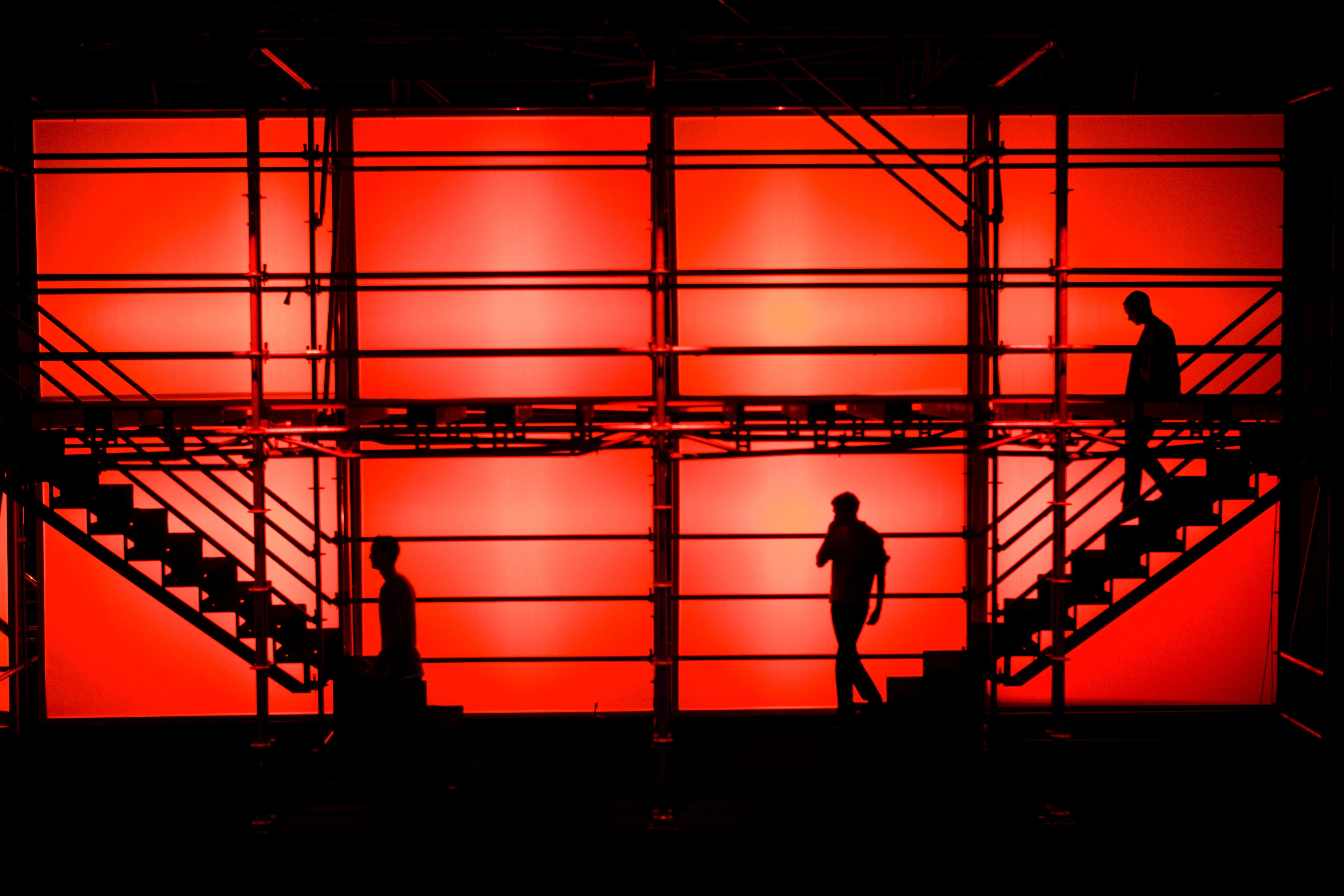 Siluetas de tres personas en un andamio con escaleras sobre un fondo rojo intenso, 080 Barcelona Fashion FW 2011-2012 producida por ESMA Events