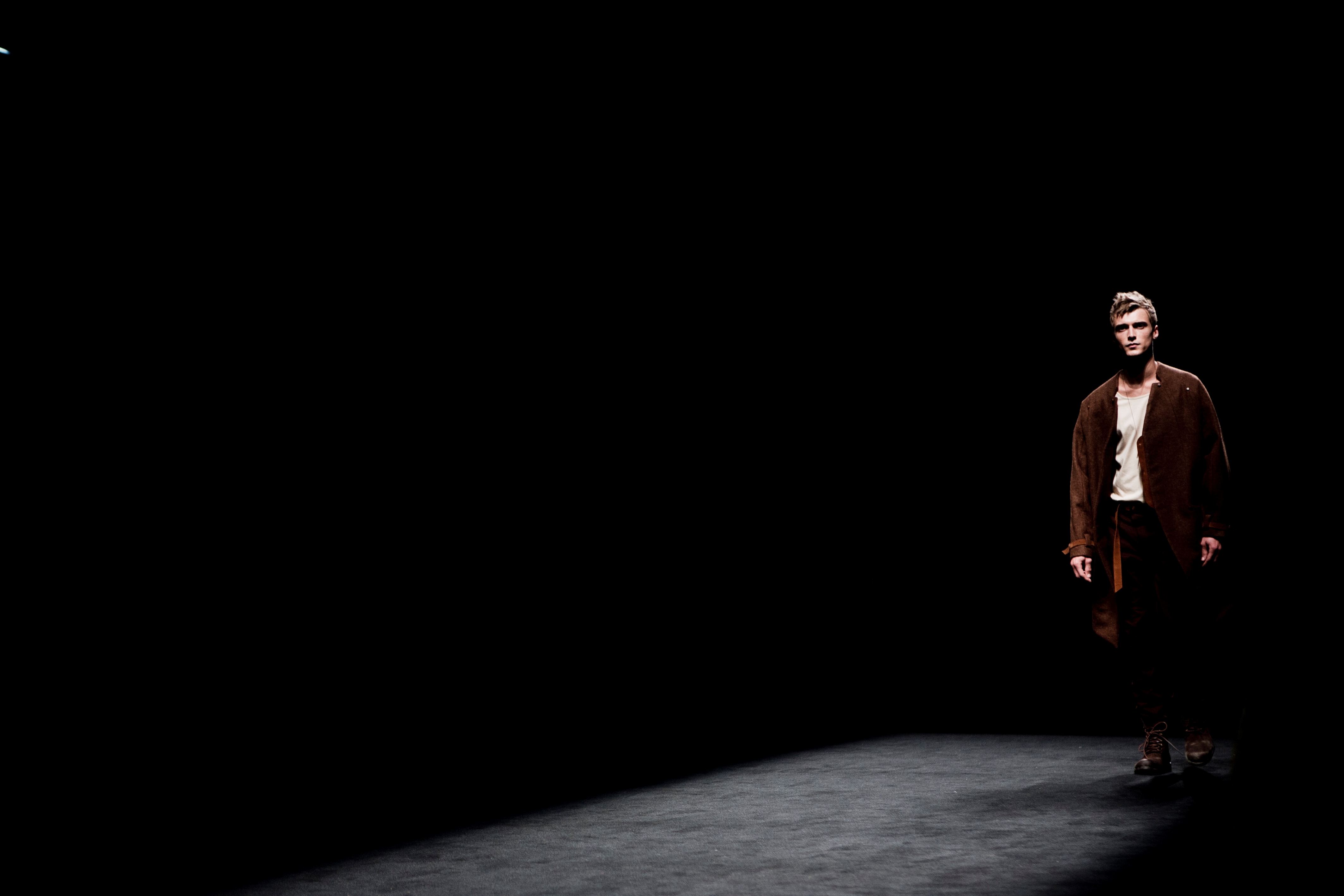 Modelo masculino con abrigo marrón avanzando en la pasarela oscura, 080 Barcelona Fashion FW 2011-2012 producida por ESMA Events