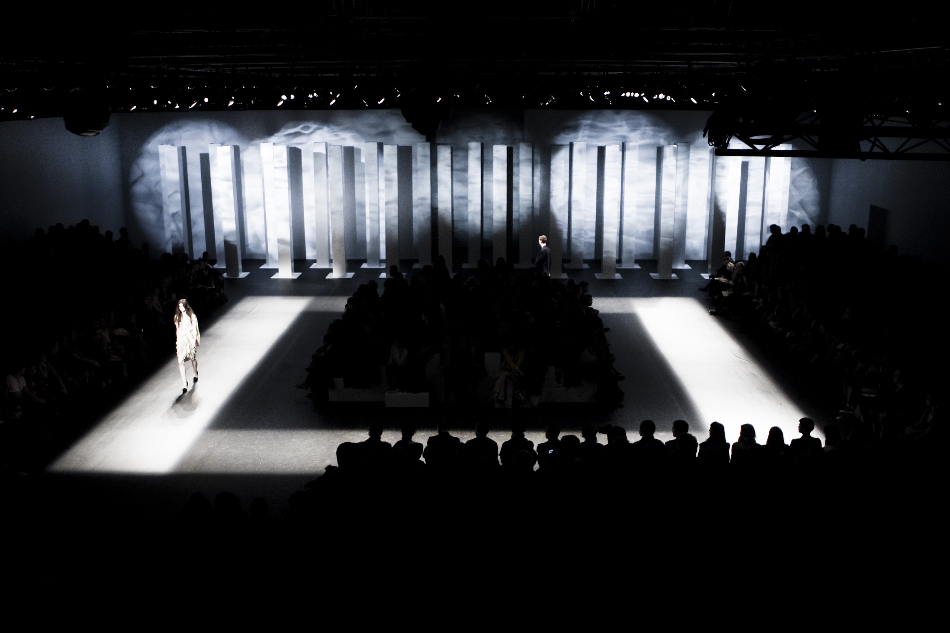 Modelo desfilando por una pasarela iluminada entre columnas blancas y público en penumbra, 080 Barcelona Fashion FW 2010-2011 producida por ESMA Events