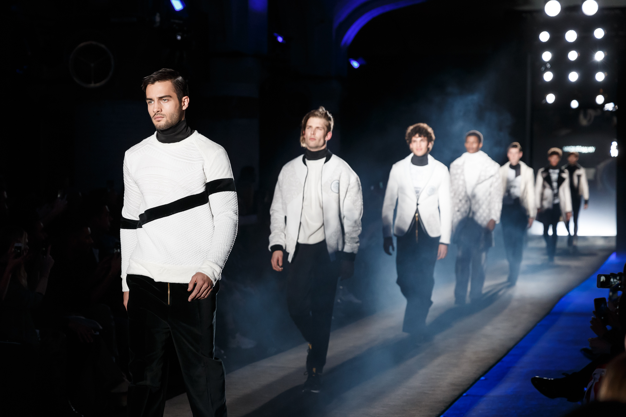 Modelos masculinos con jerséis blancos y negros desfilando sobre una pasarela azul con humo, 080 Barcelona Fashion FW 2018-2019 producida por ESMA Events