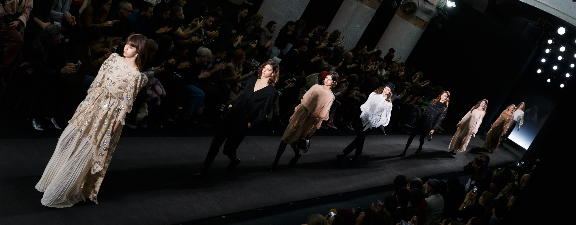 Modelos desfilando por una pasarela oscura ante el público, una con vestido claro de volantes, 080 Barcelona Fashion FW 2018-2019 producida por ESMA Events