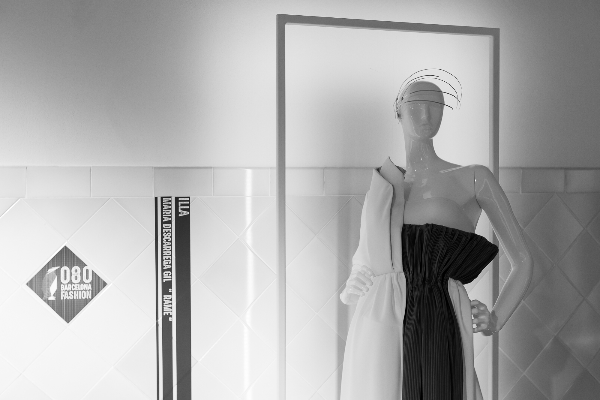 Maniquí con un vestido blanco y negro y un tocado ante una pared de azulejo con el rótulo 080, 080 Barcelona Fashion FW 2019-2020 producida por ESMA Events