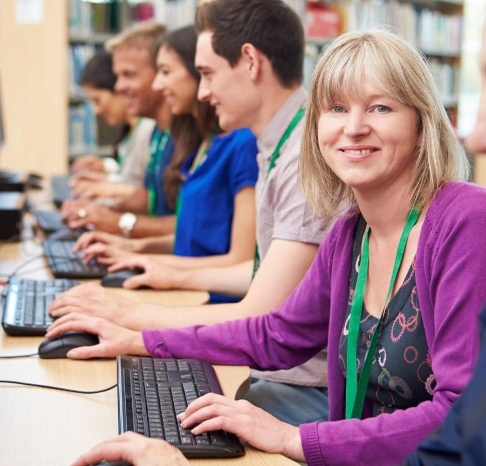 Photo of an older student looking at the camera smiling. She's sitting with 3 other college students on computer labs.