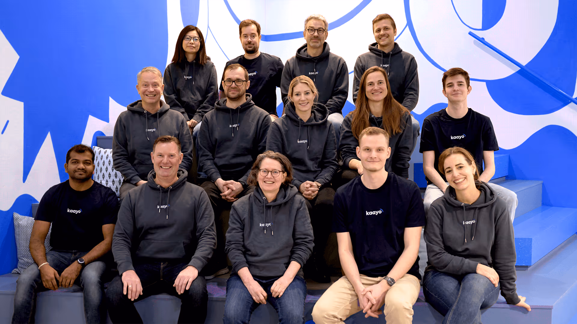 Gruppenfoto des kaayo-Teams in dunklen Kapuzenpullovern auf Bürotreppe. Fröhliche, engagierte Stimmung.