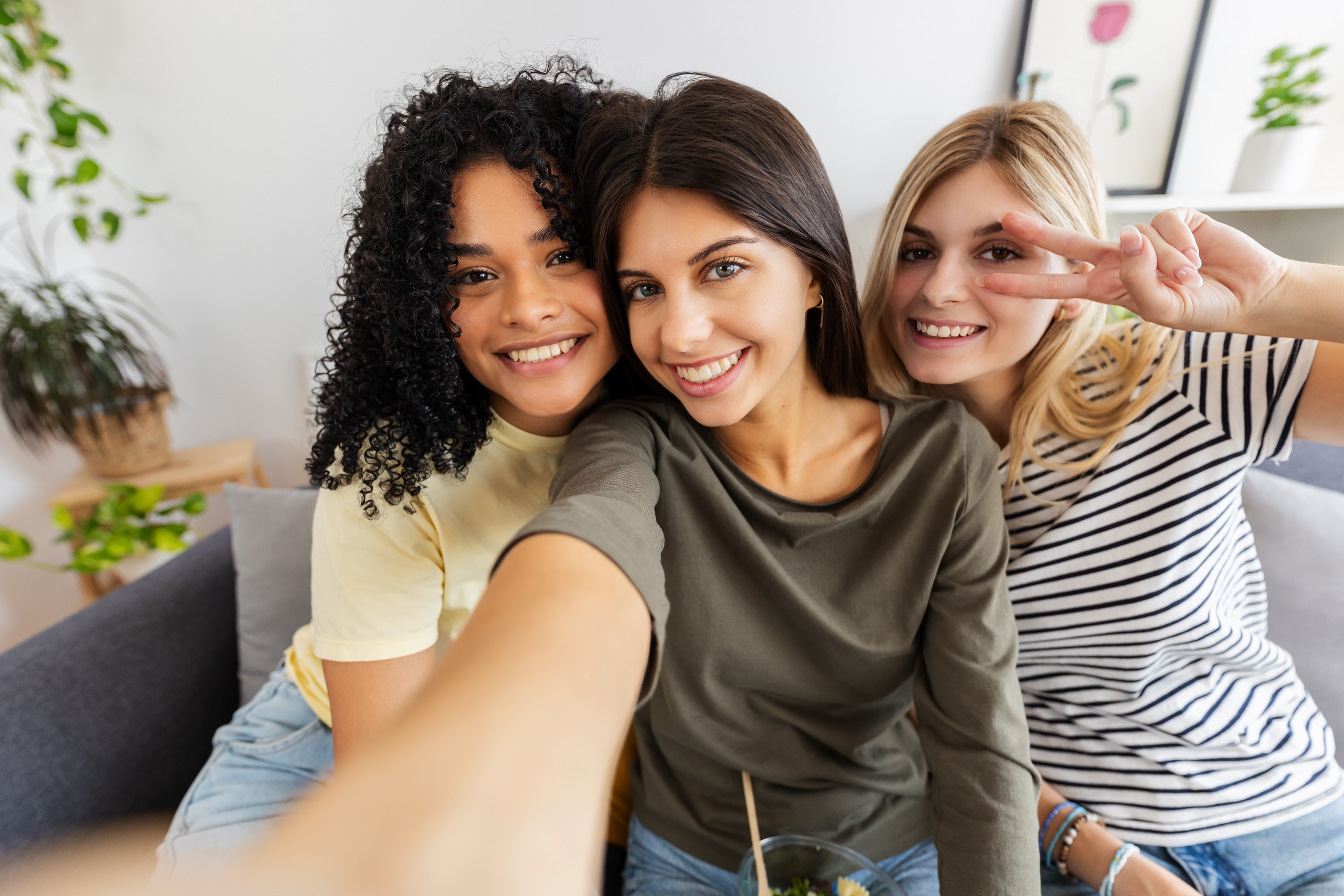 Three smiling people taking a selfie together on a gray couch in a bright living room