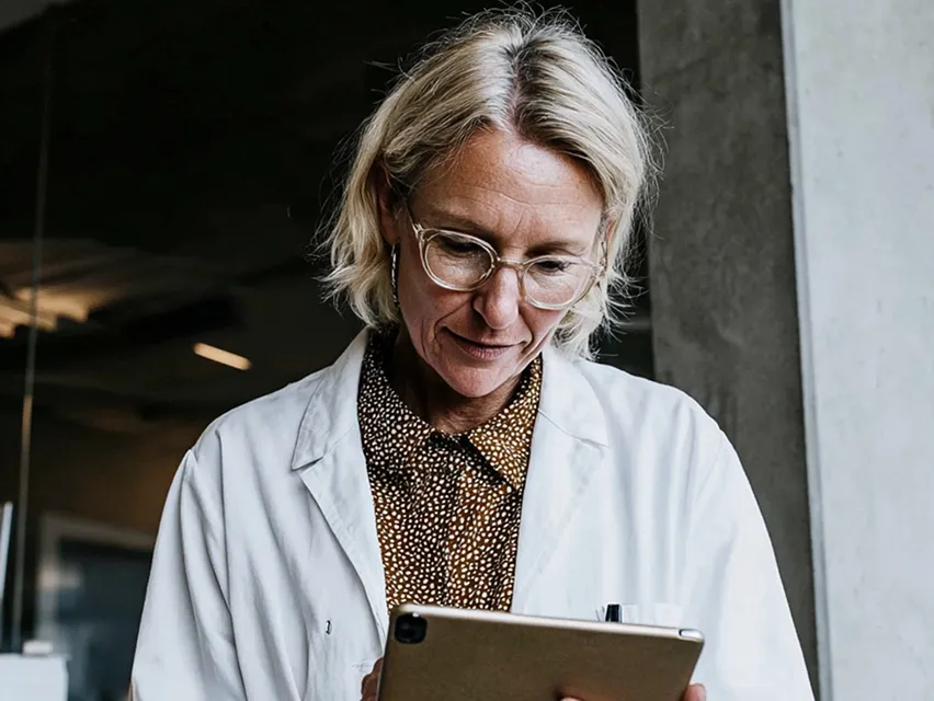 Woman with glasses wearing white coat looking at a tablet in an indoor setting.