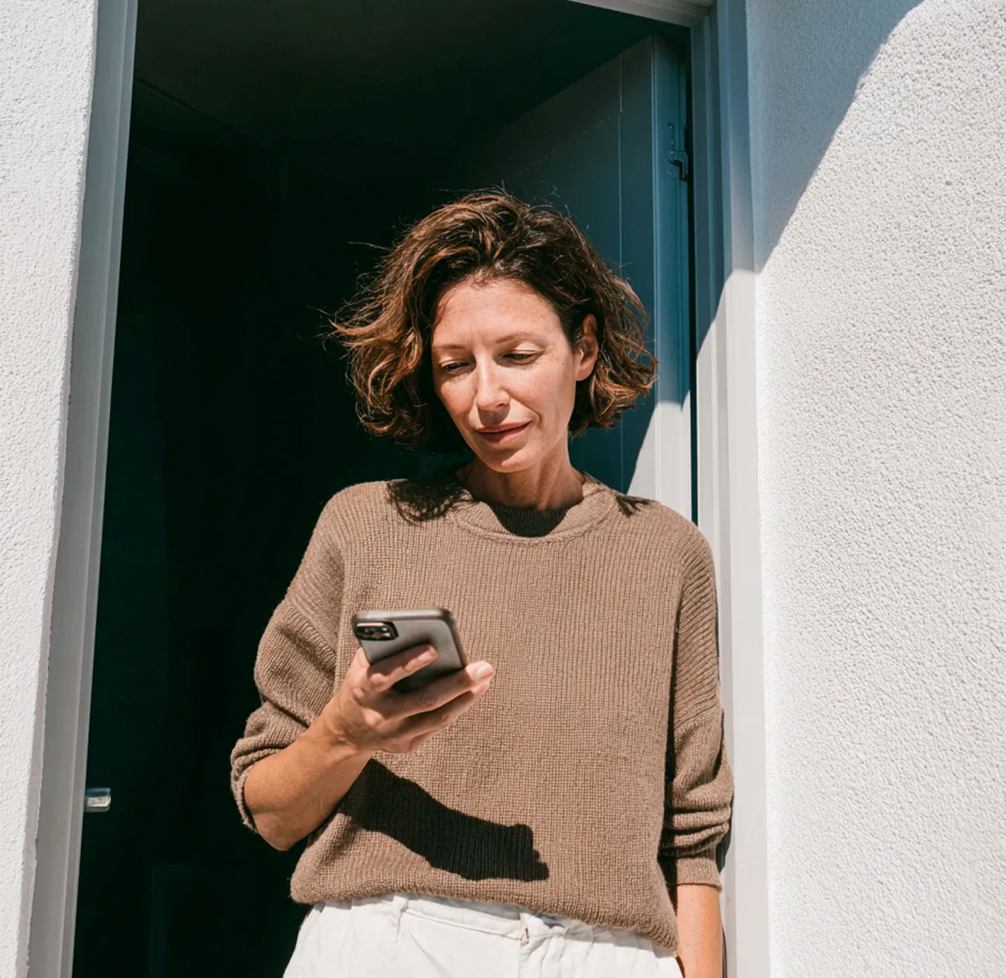 Woman in tan sweater and white pants looking at her phone while standing in a doorway with sunlight.