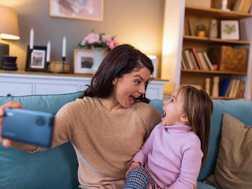 Mother and daughter sitting on a couch, making surprised faces while taking a selfie together.