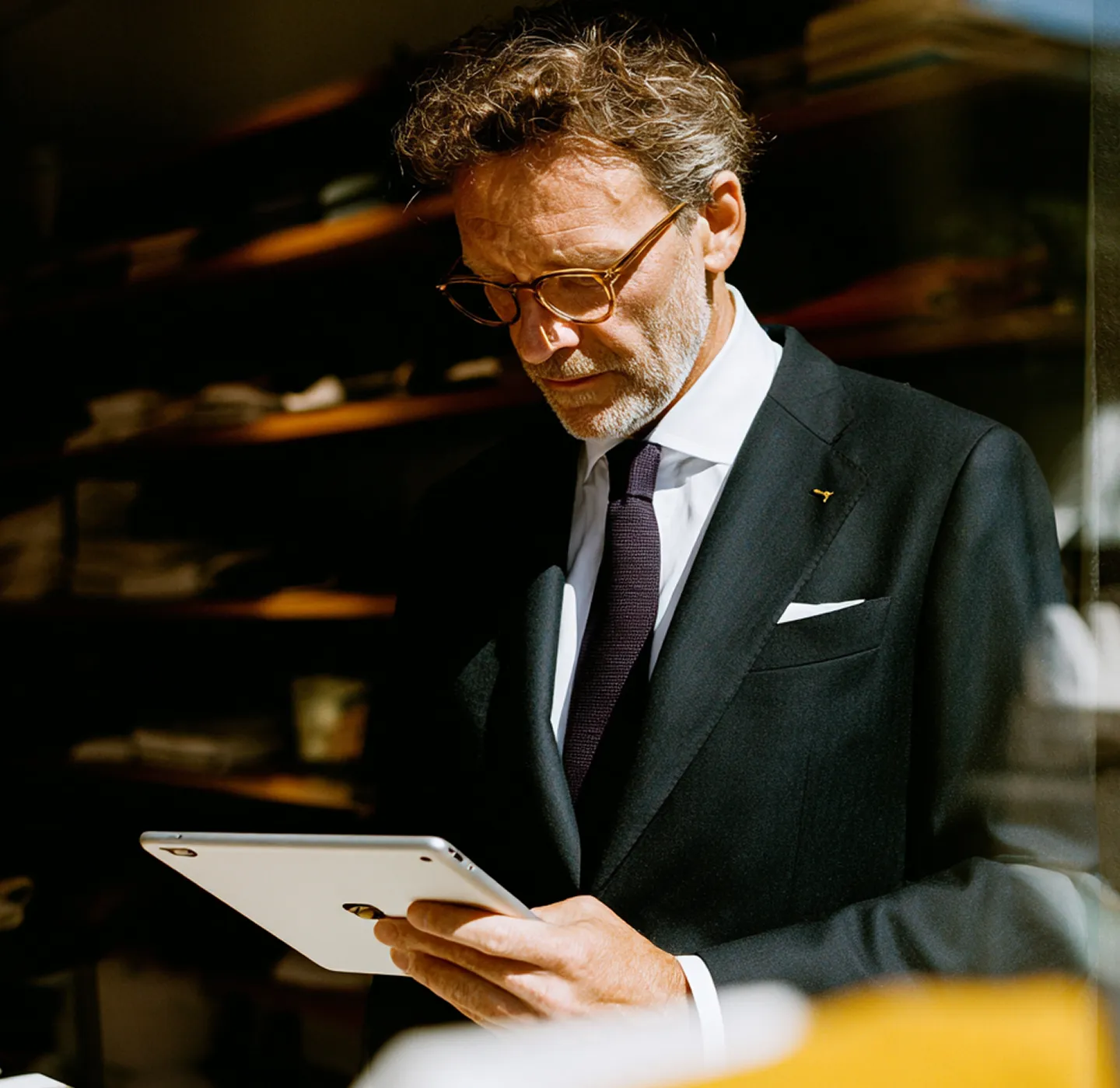 Mature man in suit reading from a tablet in a warmly lit room with shelves in the background.