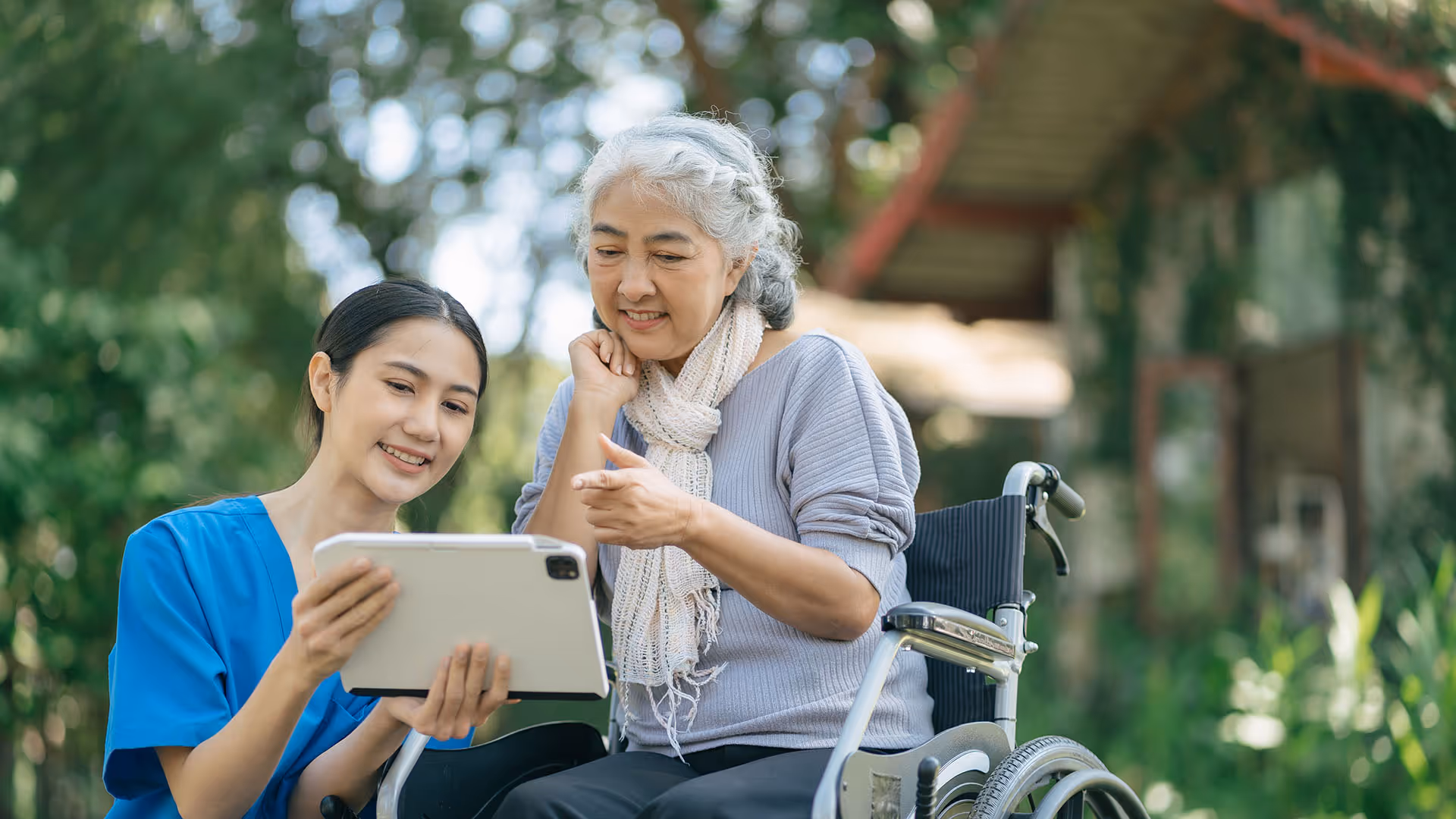 Older woman in a wheelchair talking with health aide who is holding a tablet.