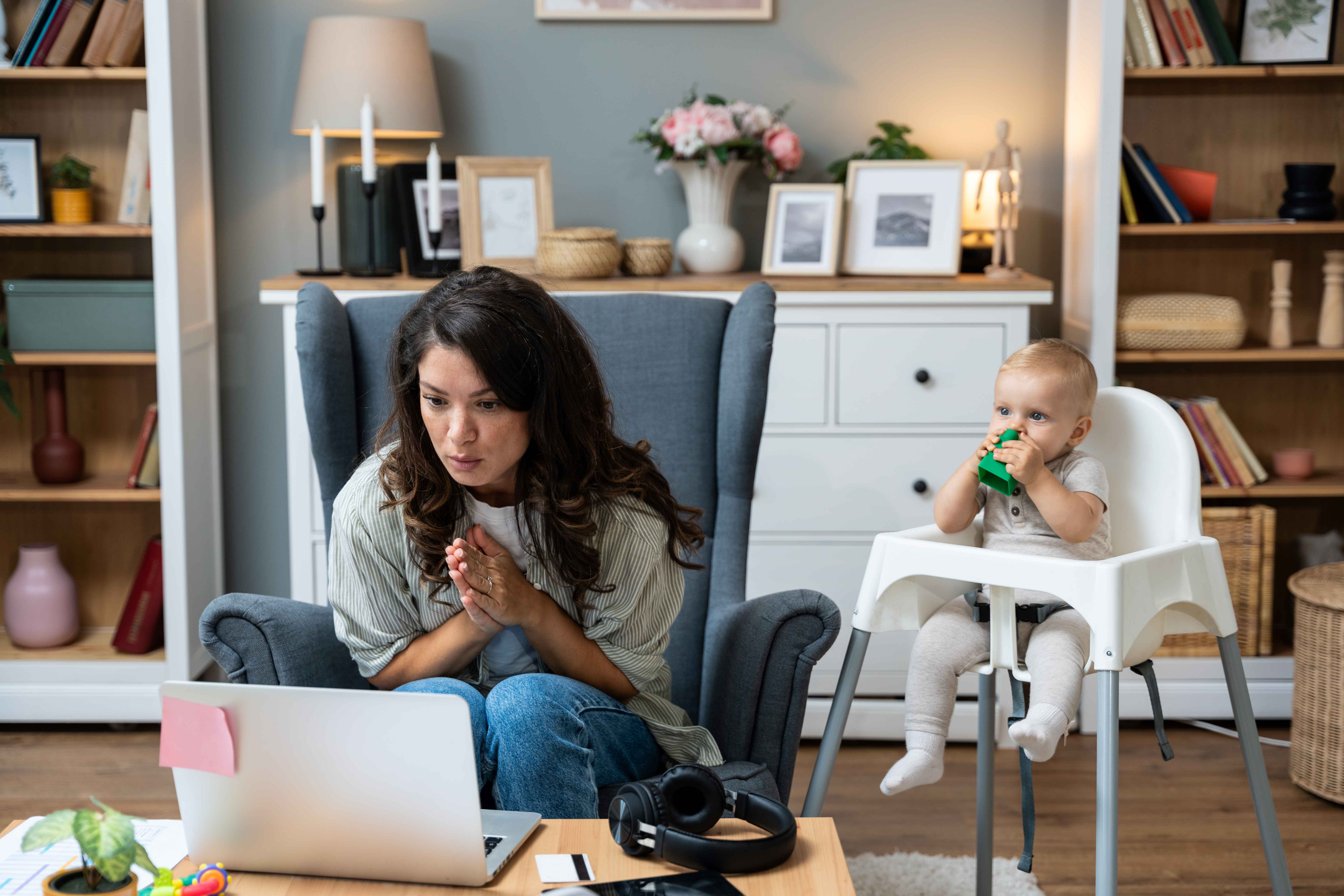 Mom uses laptop to research nanny payroll service options and household employer responsibilities while baby plays in highchair. 