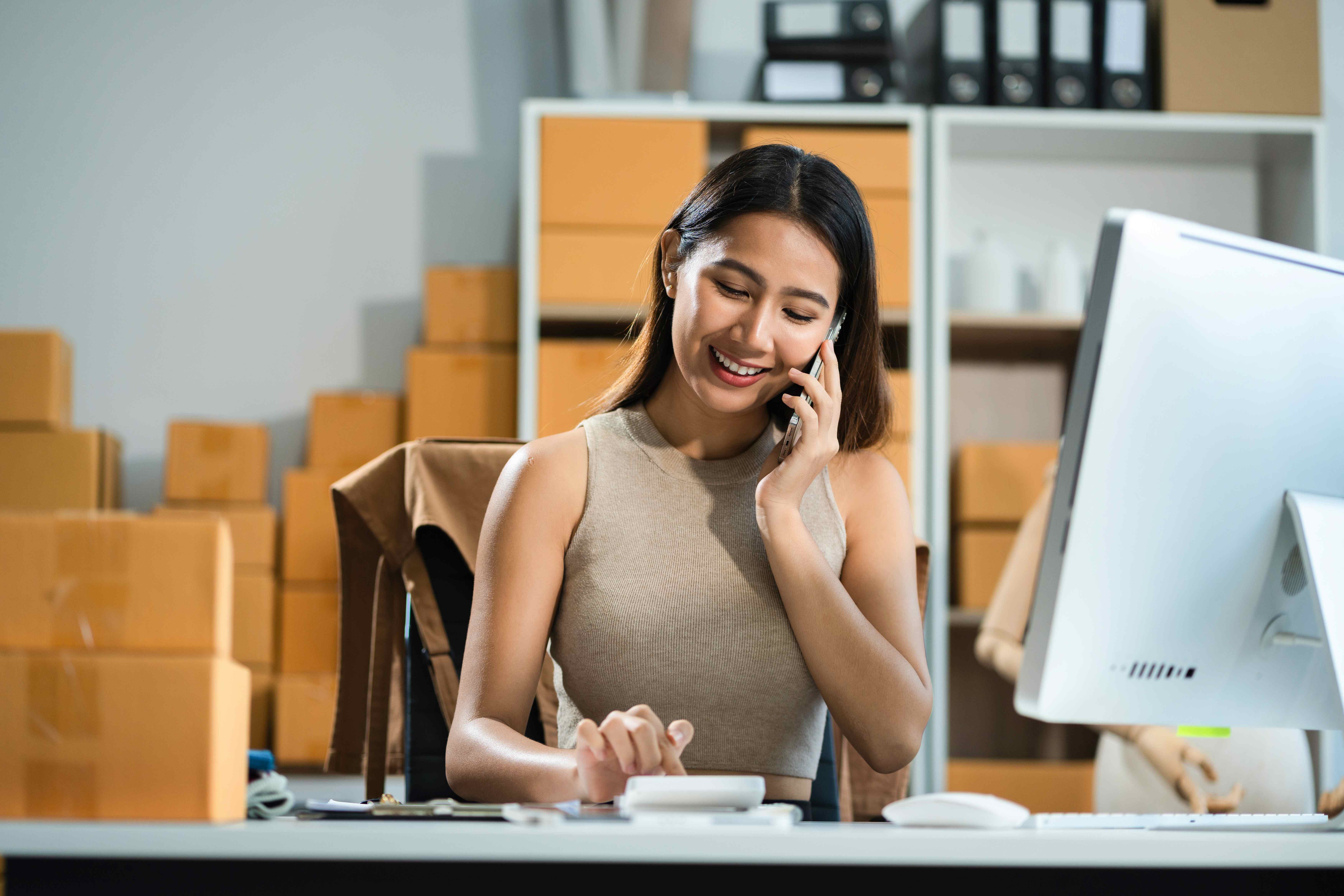 Small business owner on the phone, using a calculator and computer to figure out payroll taxes. 