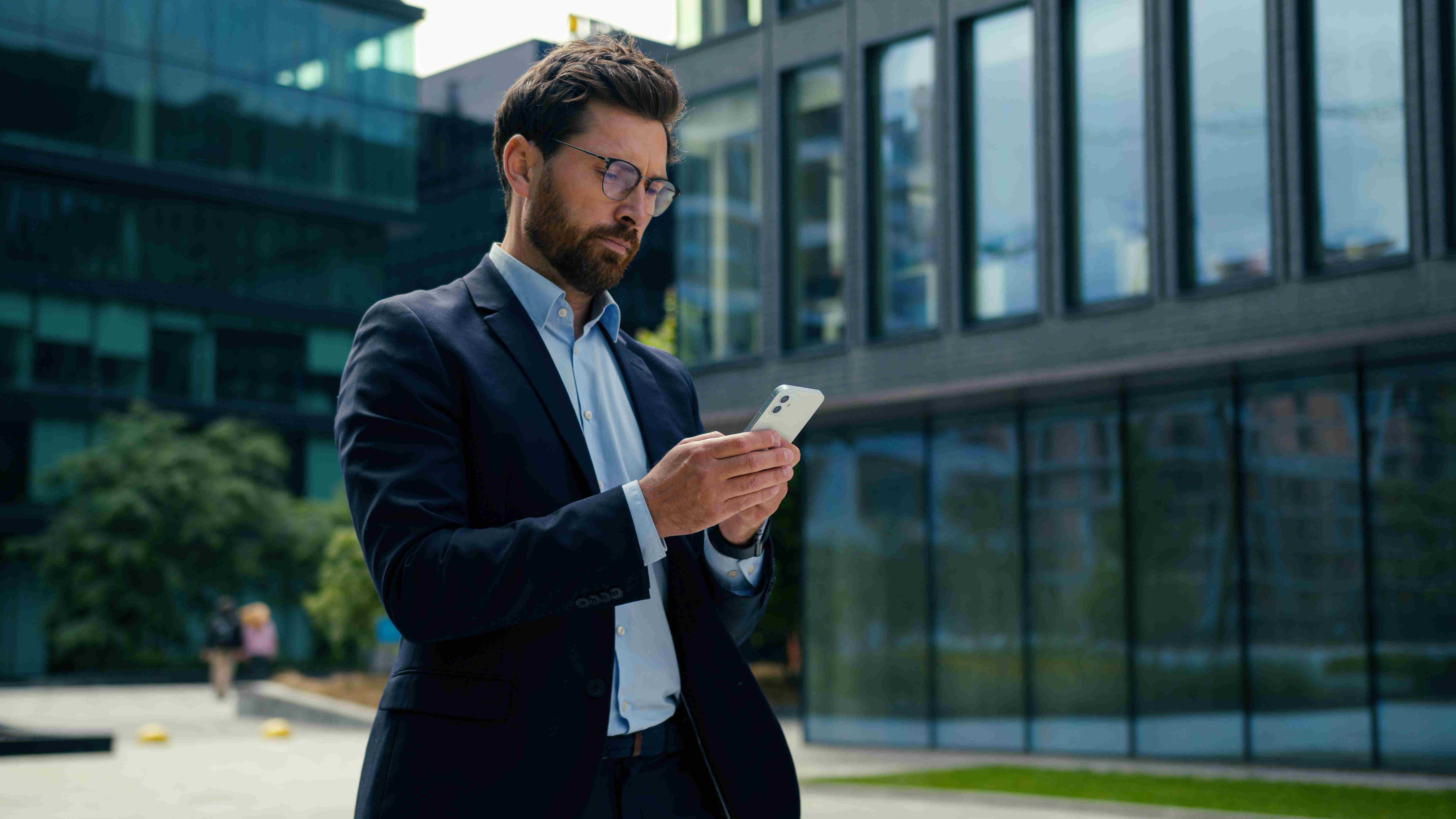 White male entrepreneur uses mobile phone outside office building to run payroll. 