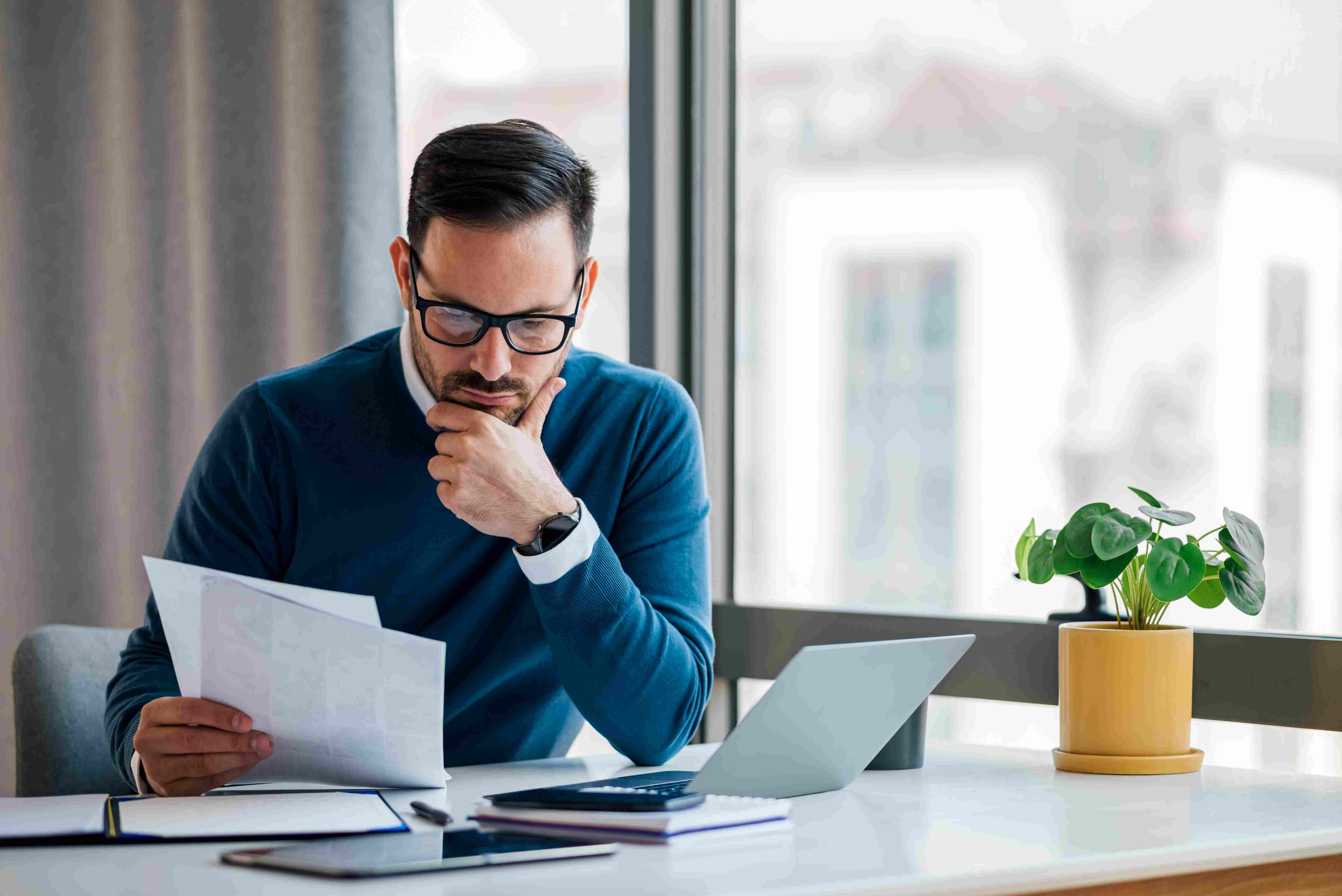 Male S-corp owner reviews papers to set up s corp payroll using laptop.