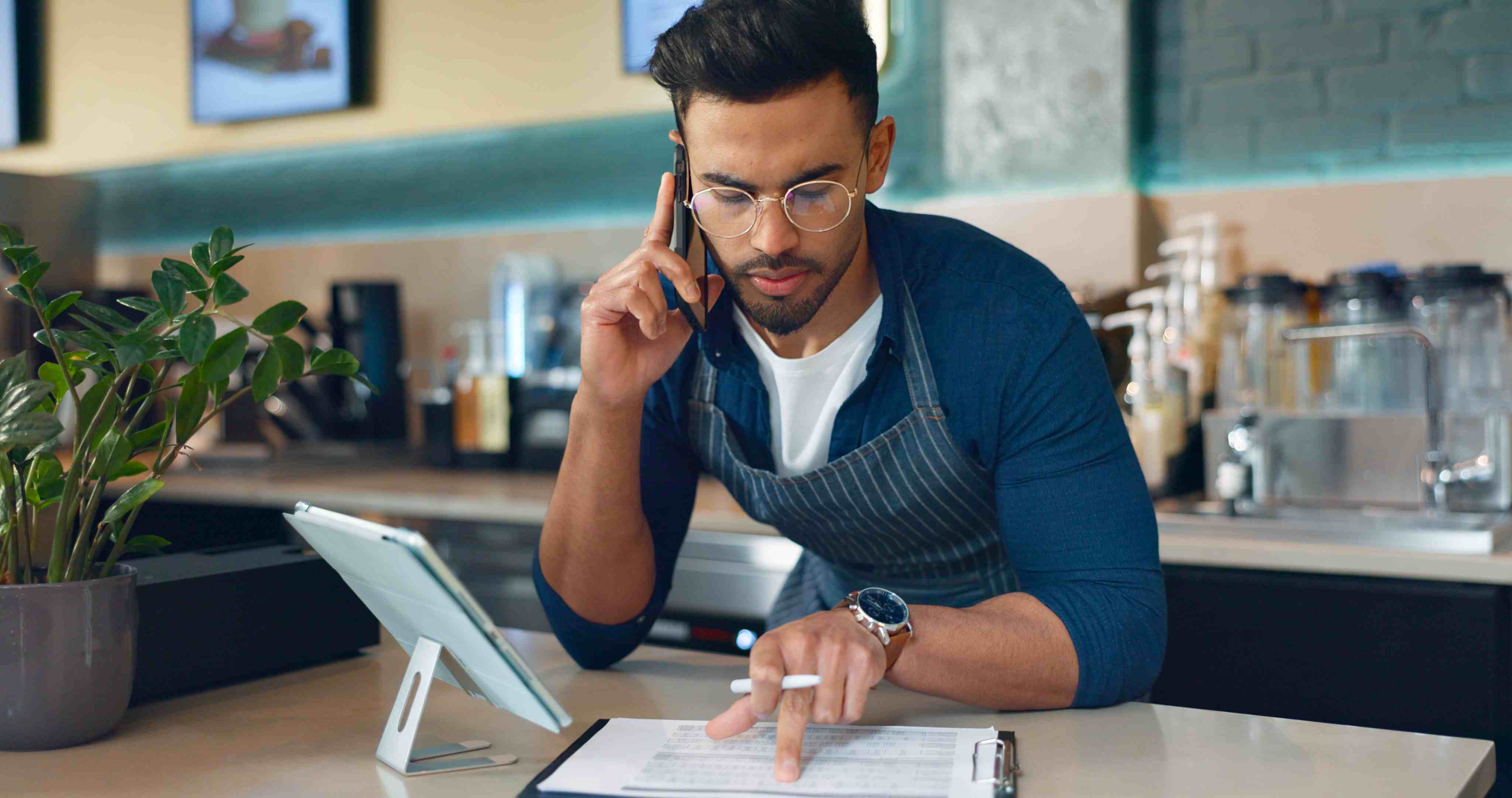 Coffee shop owner reviews payroll software at counter with tablet and clipboard