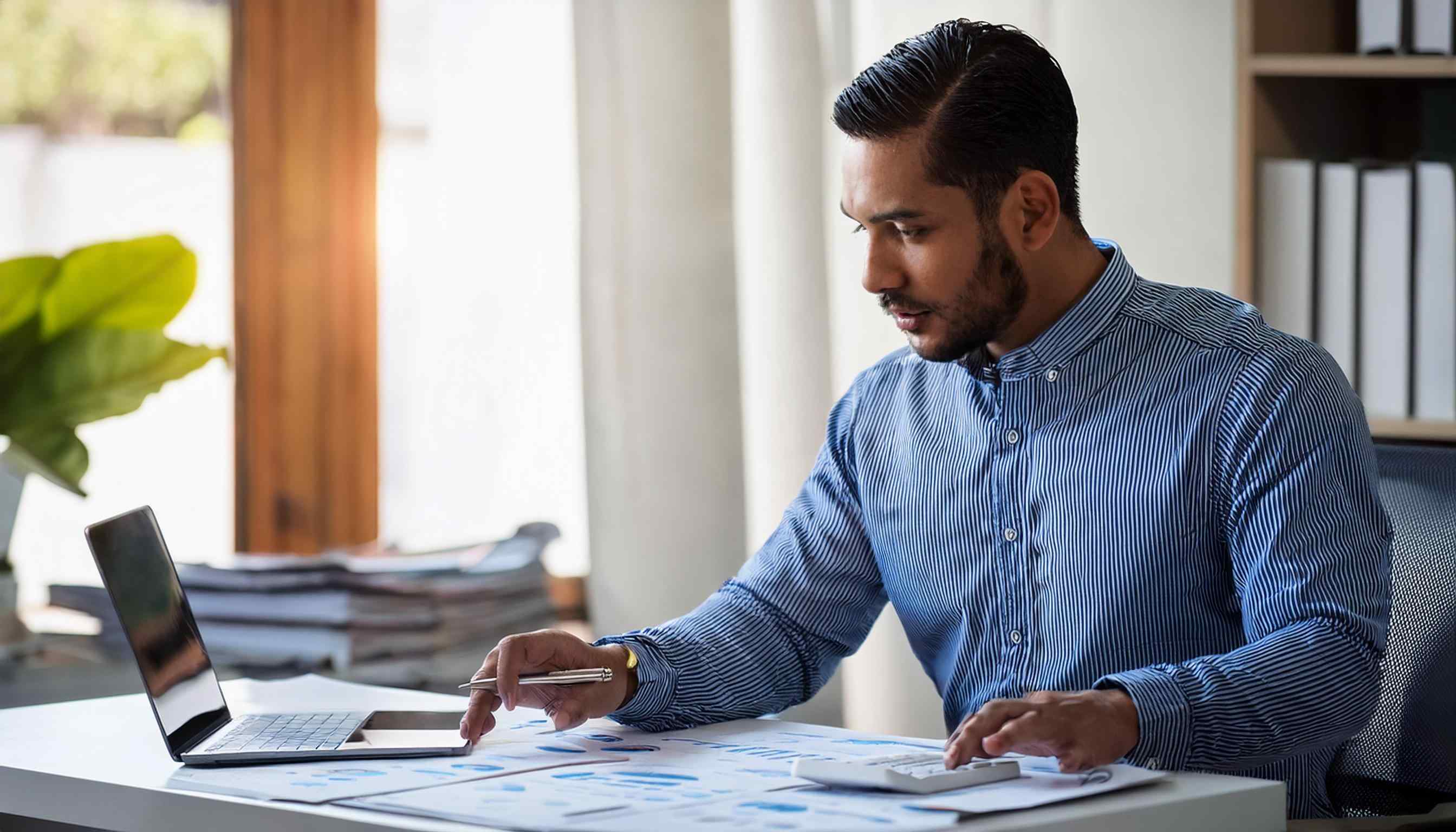 Financial professional at desk with laptop, calculator, paperwork.