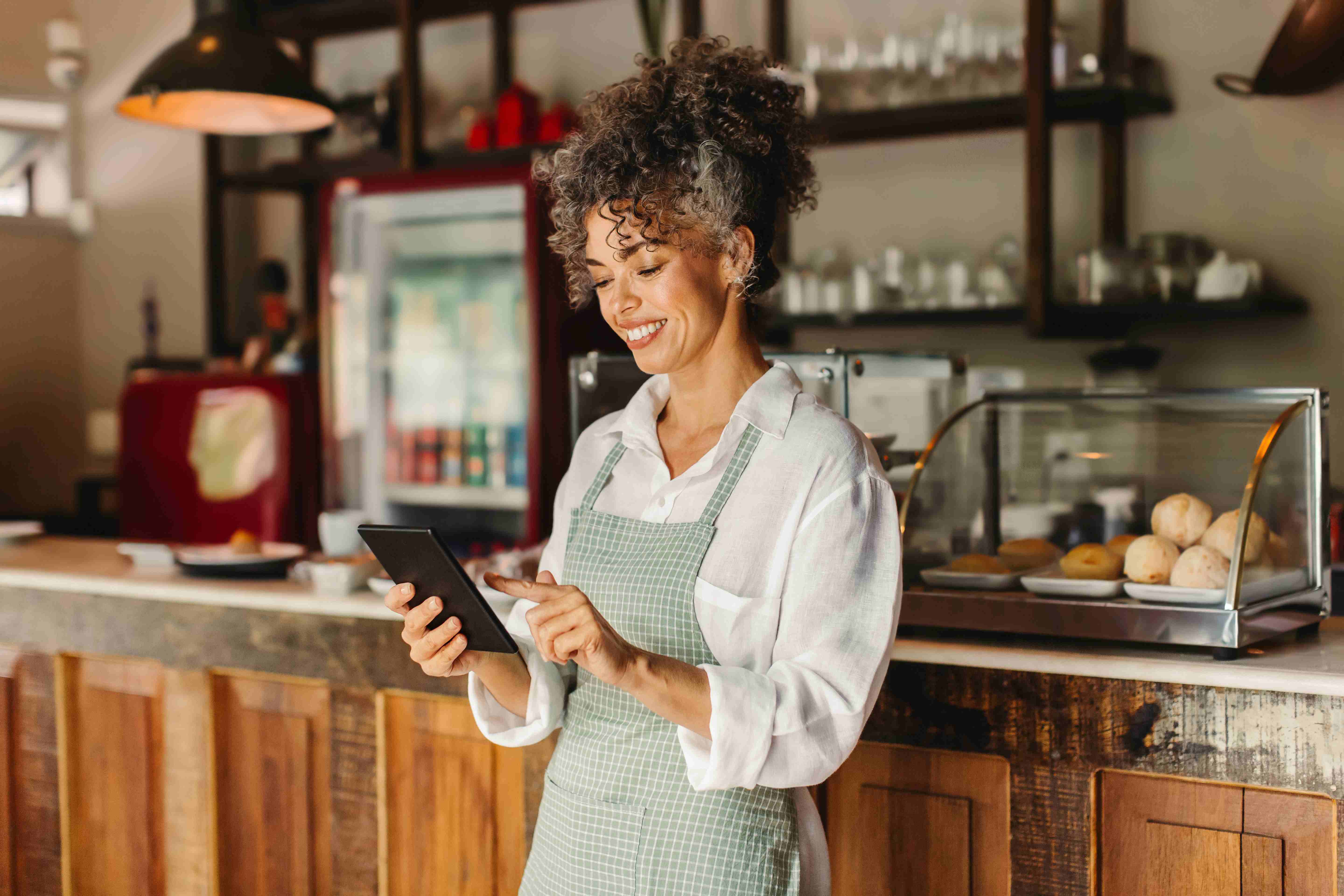 Cafe owner stands in front of counter using tablet. 