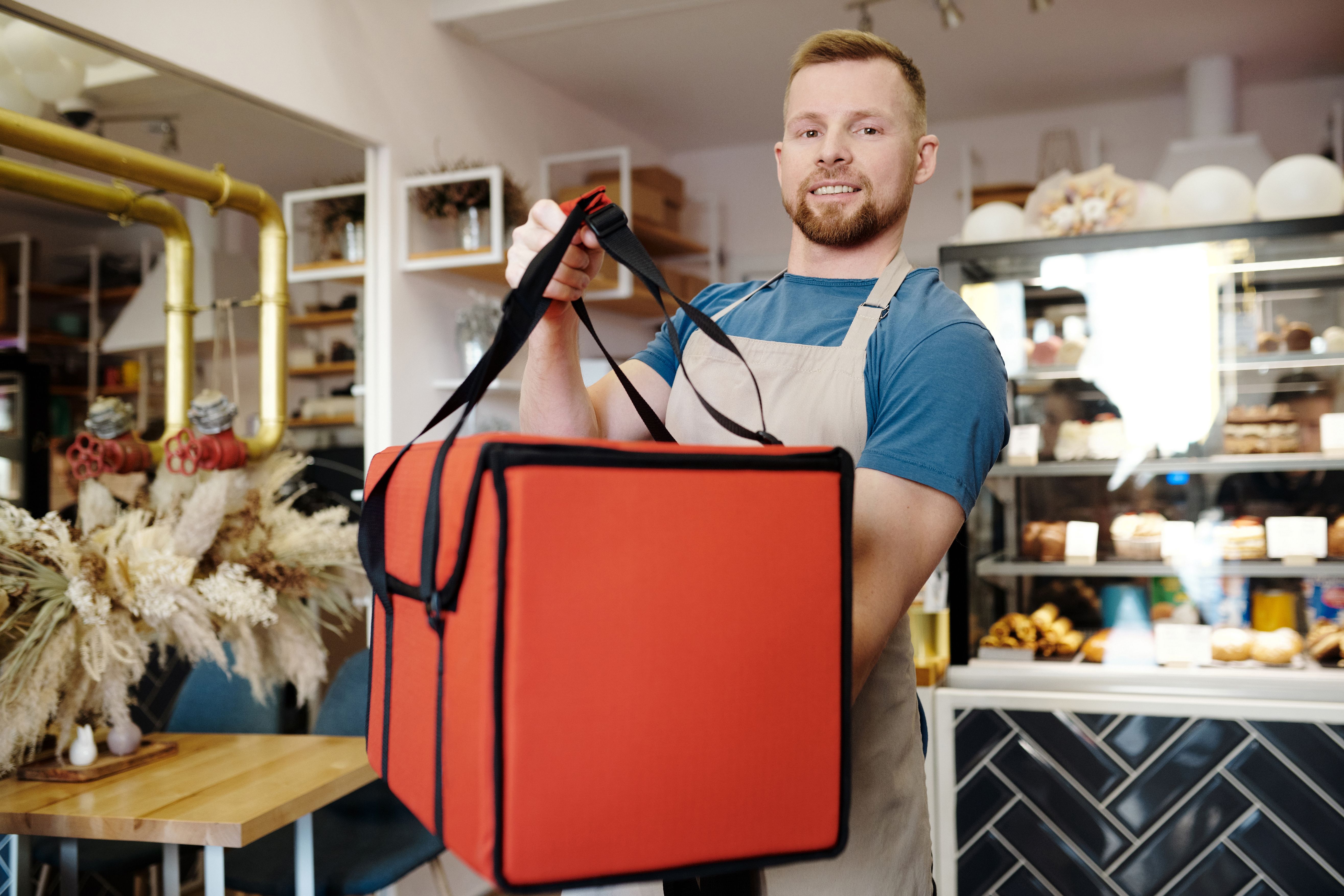 A man in a blue shirt holds up a red, industrial delivery cooler.