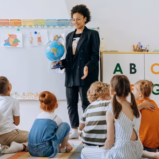 A small group of preschool-aged children sit on the floor, listening to a female teacher in a classroom.