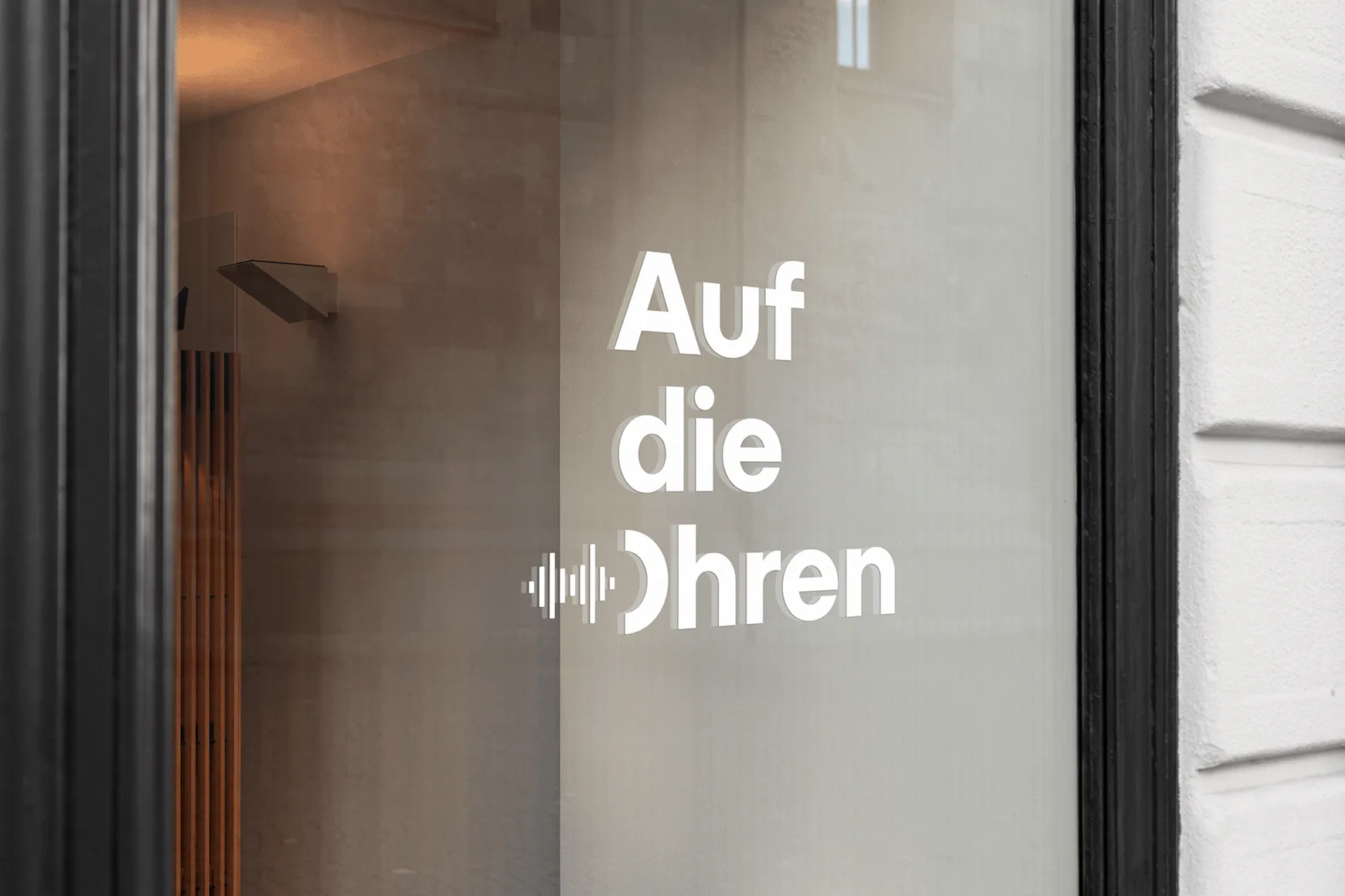 White three-line word mark of the company "Auf die Ohren" on a window in a modern sans-serif font. An abstract, reduced audio wave runs over into the "O" of the company name.