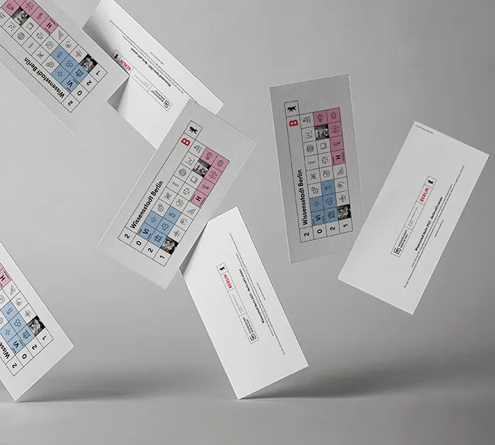 Several falling flyers of the Senate in Berlin with the current theme for the annual card 2021 with the motto " City of Knowledge Berlin". The design is a geometric illustration in the colors white, black, red, light blue and pink, structured like a periodic table filled with various icons on the subject of science.