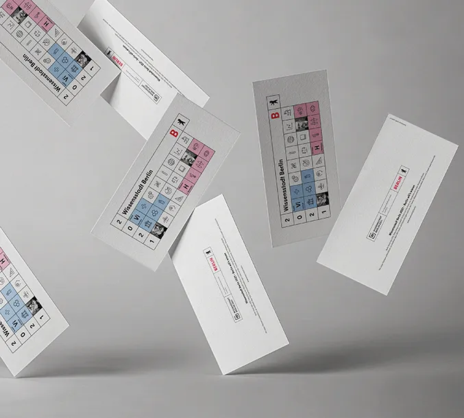 Several falling flyers of the Senate in Berlin with the current theme for the annual card 2021 with the motto " City of Knowledge Berlin". The design is a geometric illustration in the colors white, black, red, light blue and pink, structured like a periodic table filled with various icons on the subject of science.