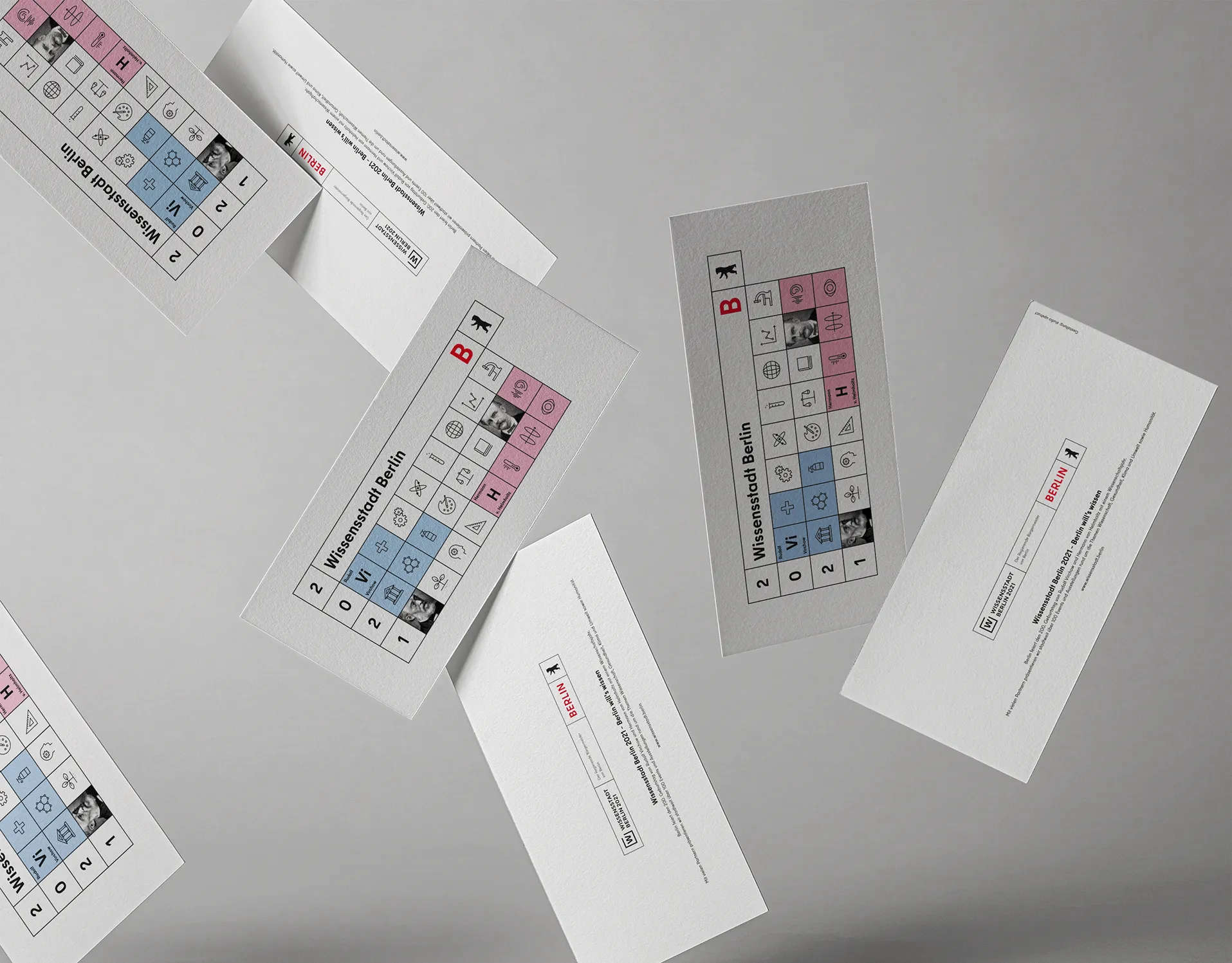 Several falling flyers of the Senate in Berlin with the current theme for the annual card 2021 with the motto " City of Knowledge Berlin". The design is a geometric illustration in the colors white, black, red, light blue and pink, structured like a periodic table filled with various icons on the subject of science.