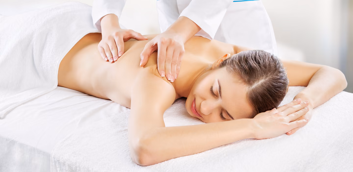 A woman lies face down on a massage table with eyes closed while a therapist's hands massage her upper back and shoulders.