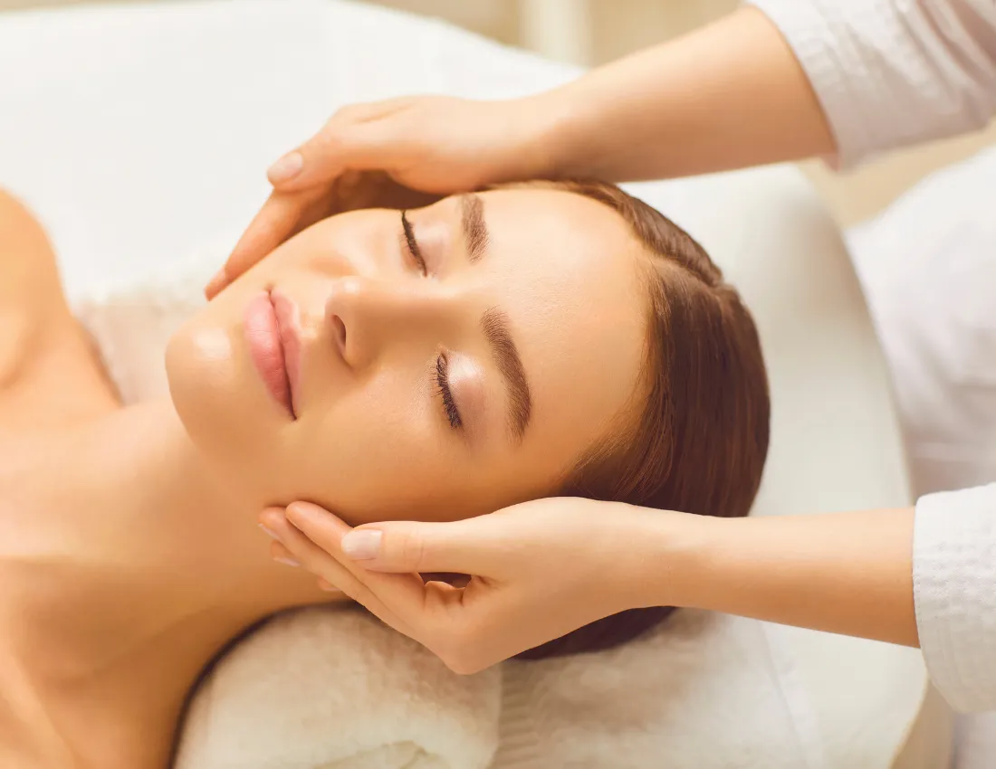 Close-up of a woman with closed eyes receiving a gentle face massage from two hands while lying on a towel in a spa setting.