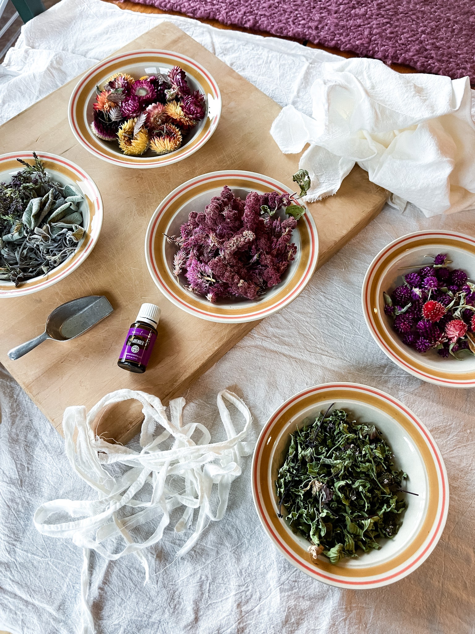 Dried flowers and herbs in bowls on a cutting board with lavender essential oil and flour sack towels-ready to make DIY sachets.