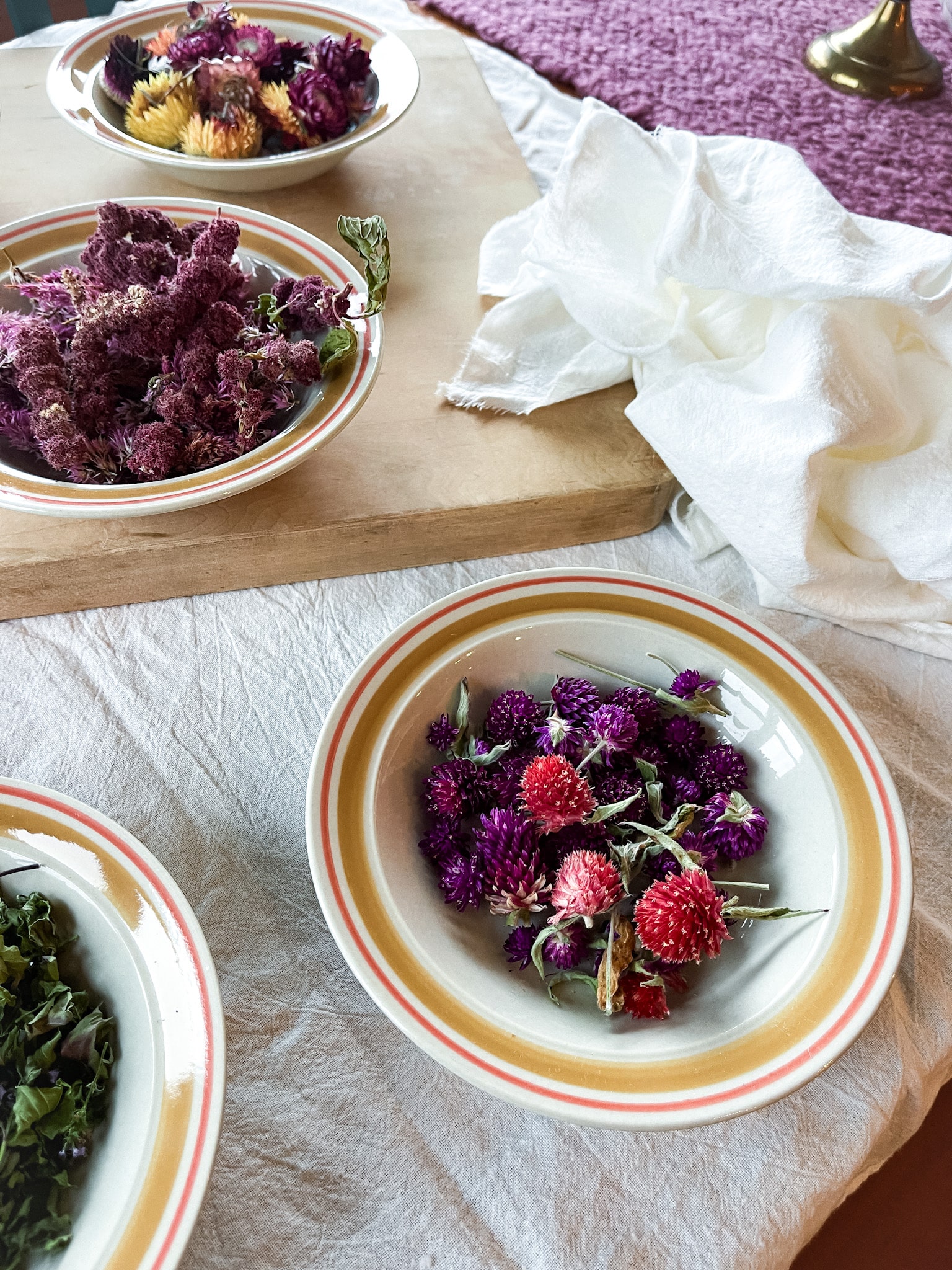 Dried flowers and herbs in bowls on a cutting board with lavender essential oil and flour sack towels-ready to make DIY sachets.
