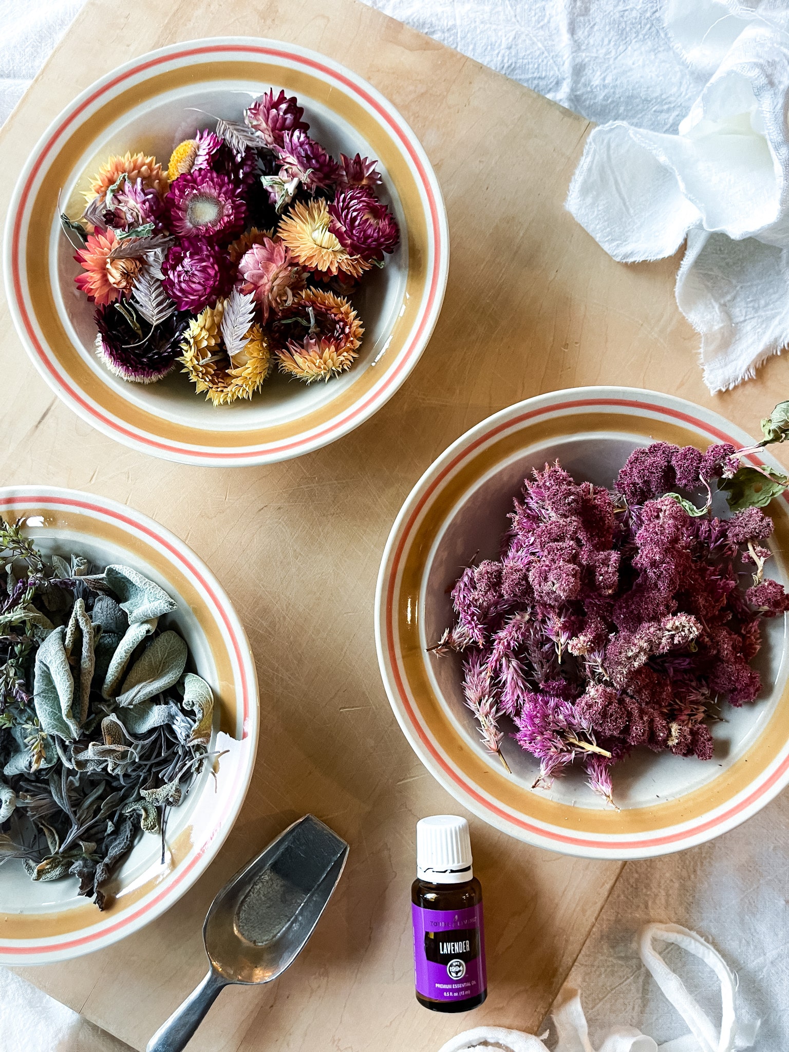 Dried flowers and herbs in bowls on a cutting board with lavender essential oil and flour sack towels-ready to make DIY sachets.