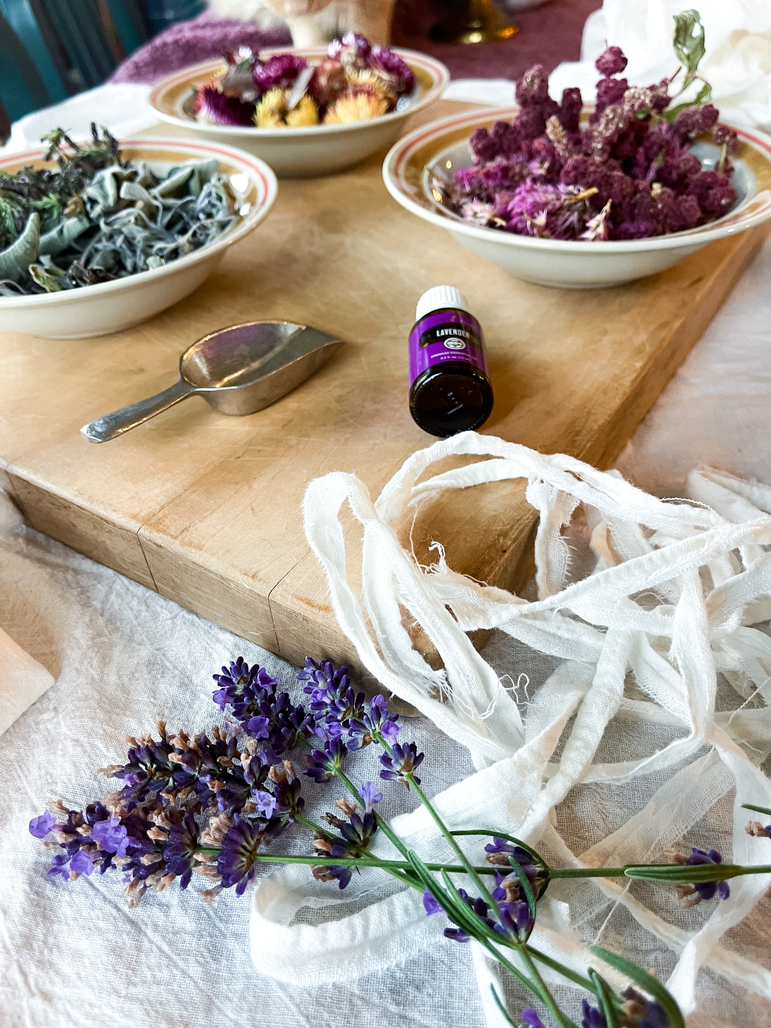 Dried flowers and herbs in bowls on a cutting board with lavender essential oil and flour sack towels-ready to make DIY sachets.