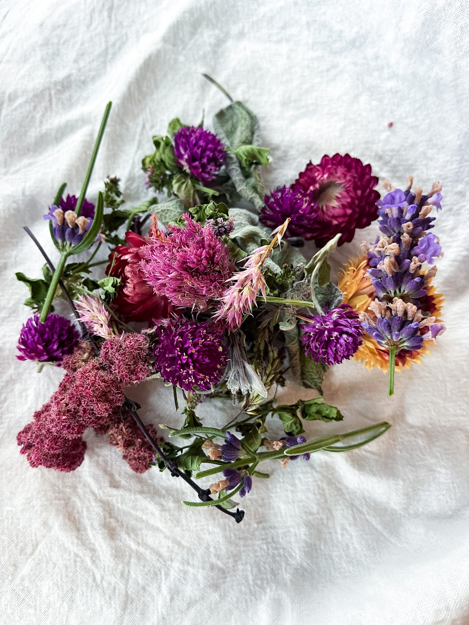 Dried flowers and herbs in bowls on a cutting board with lavender essential oil and flour sack towels-ready to make DIY sachets.