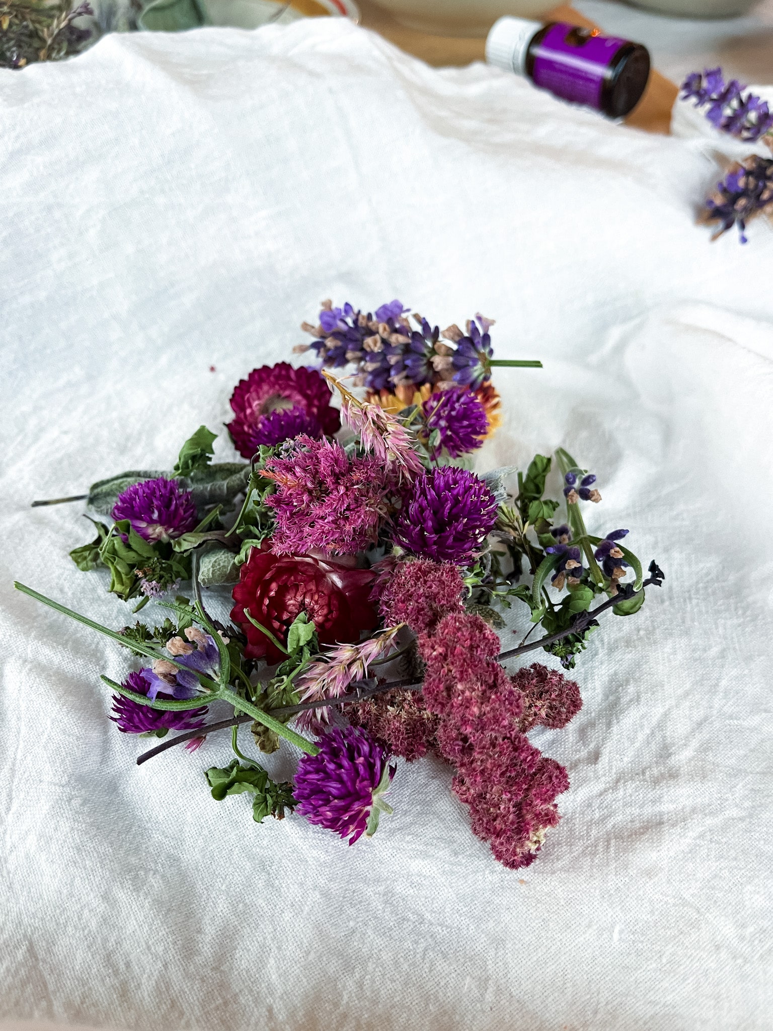 Dried flowers and herbs in bowls on a cutting board with lavender essential oil and flour sack towels-ready to make DIY sachets.