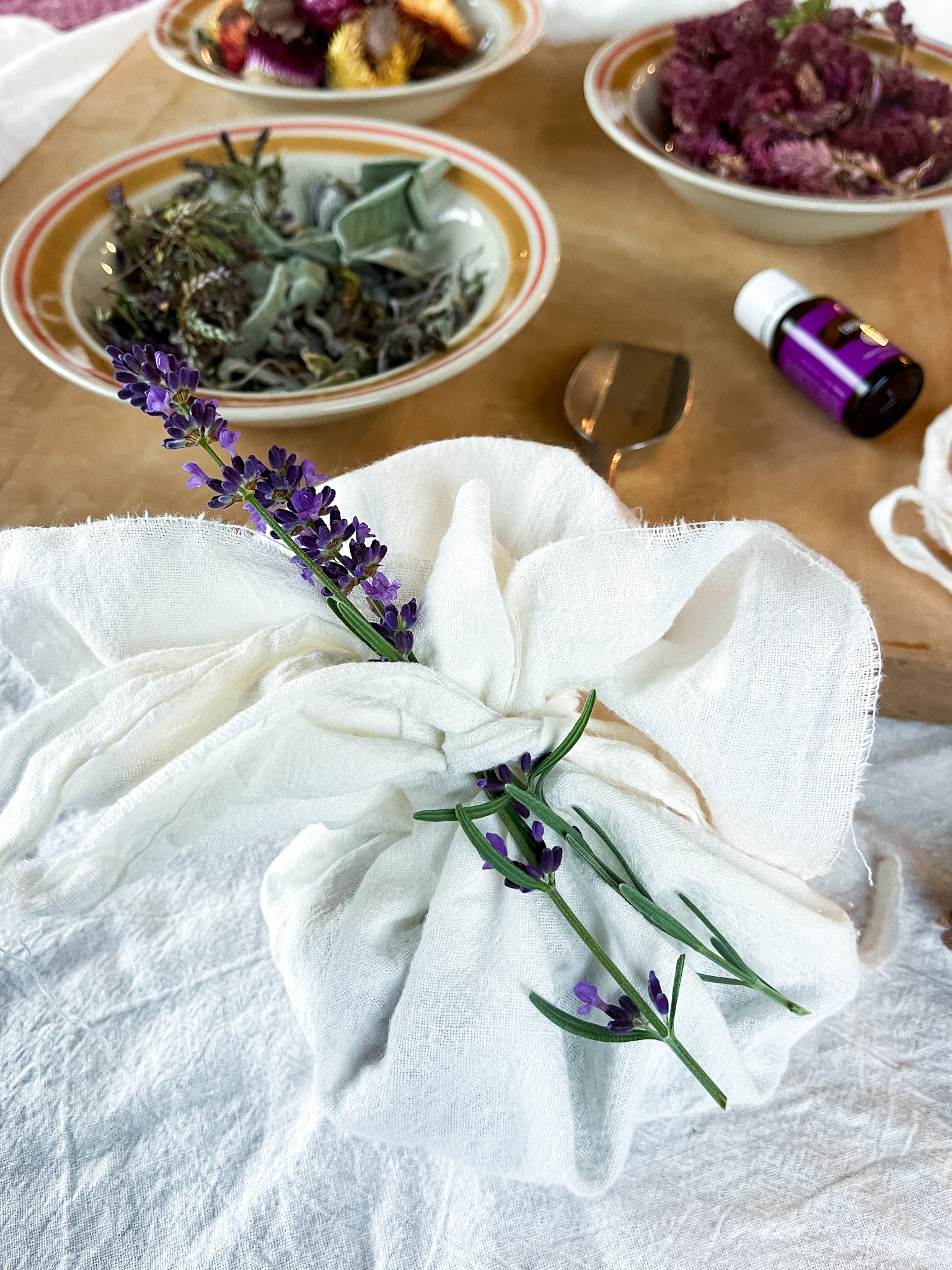 Dried flowers and herbs in bowls on a cutting board with lavender essential oil and flour sack towels-ready to make DIY sachets.