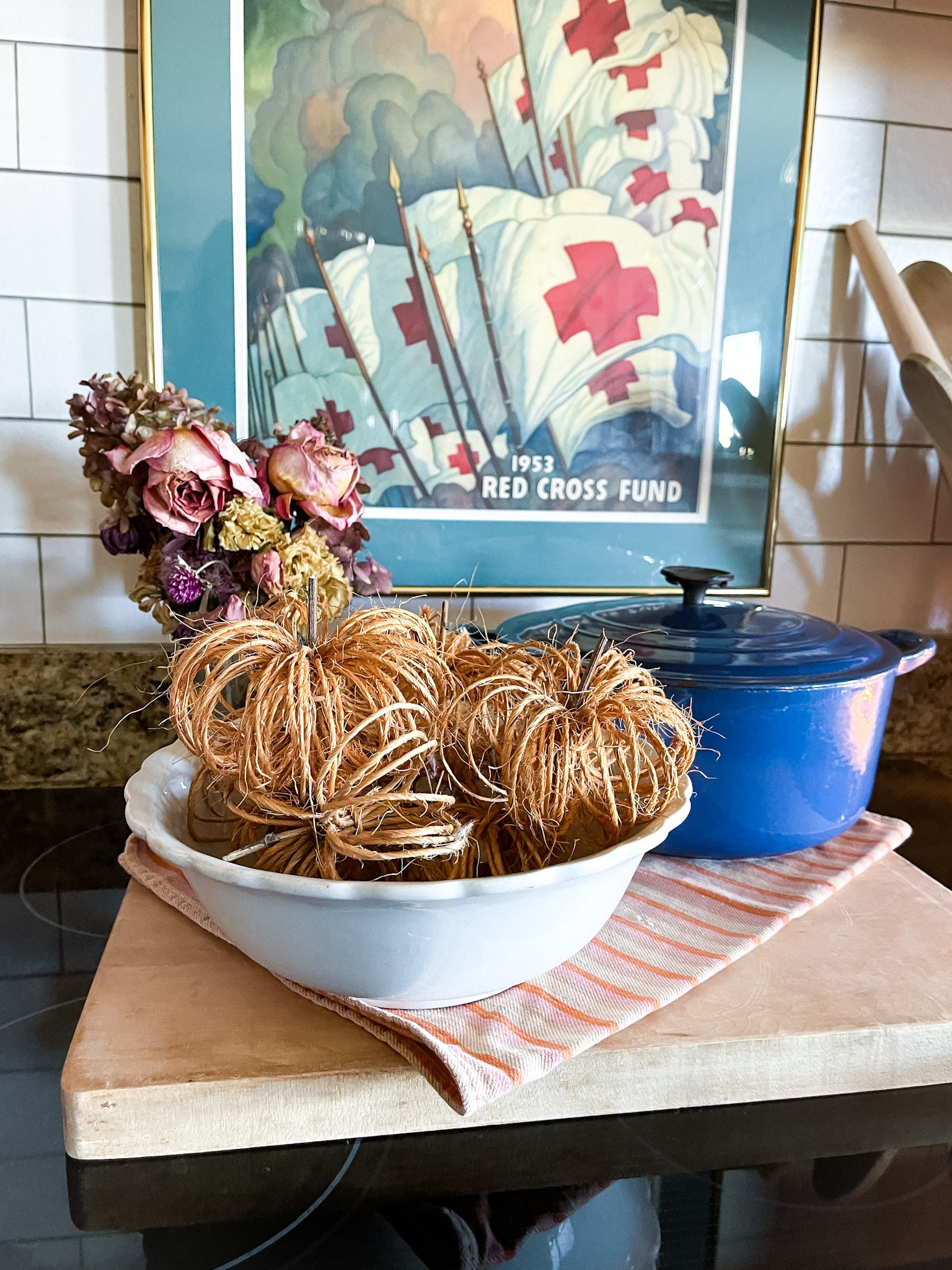 twine pumpkins in an ironstone bowl on a cutting board with a bouquet of dried flowers and a blue le creuset dutch oven