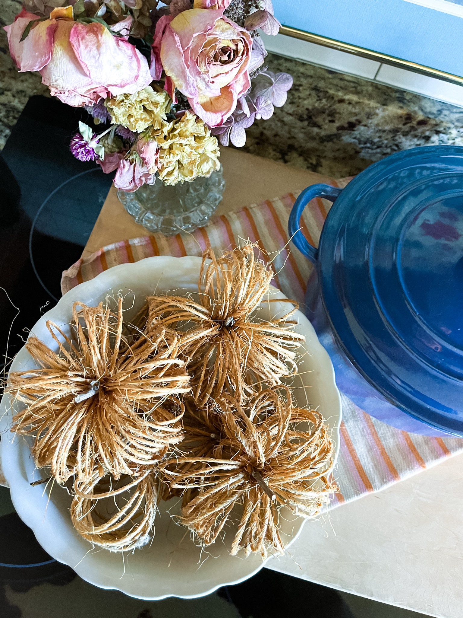 twine pumpkins in an ironstone bowl on a cutting board with a bouquet of dried flowers and a blue le creuset dutch oven