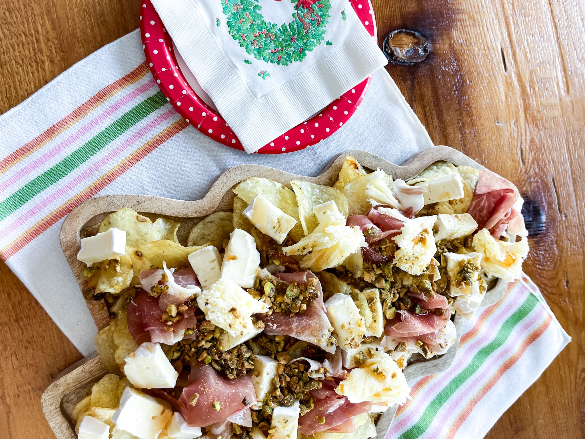 Tree shaped serving board with chipcuterie on it, sitting on a farmhouse table on a towel with vintage plates and napkins. Ingredients from Trader Joe's: potato chips, brie, prosciutto, pistachios, hot honey, tete de moine rosettes