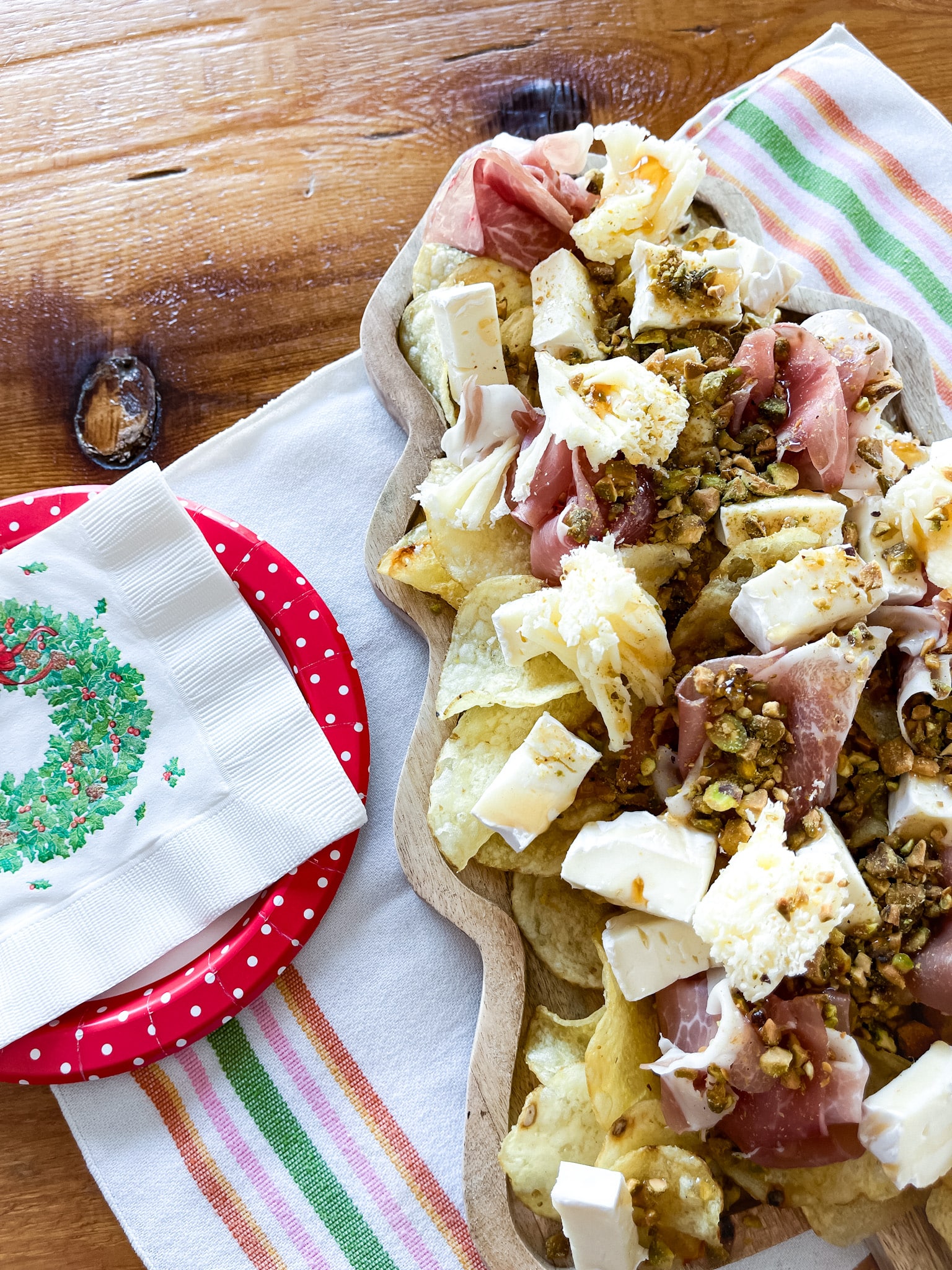 Tree shaped serving board with chipcuterie on it, sitting on a farmhouse table on a towel with vintage plates and napkins. Ingredients from Trader Joe's: potato chips, brie, prosciutto, pistachios, hot honey, tete de moine rosettes