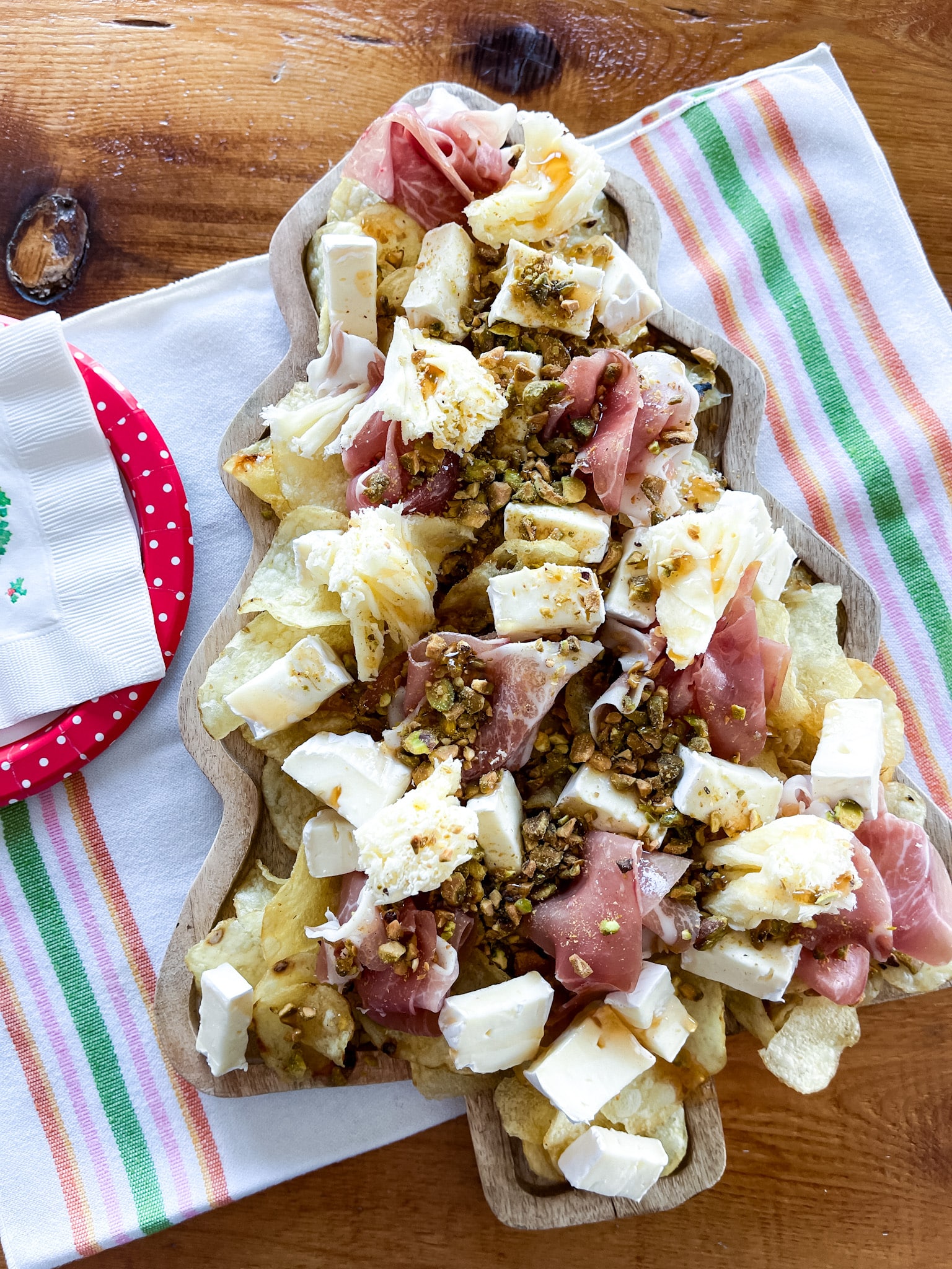 Tree shaped serving board with chipcuterie on it, sitting on a farmhouse table on a towel with vintage plates and napkins. Ingredients from Trader Joe's: potato chips, brie, prosciutto, pistachios, hot honey, tete de moine rosettes