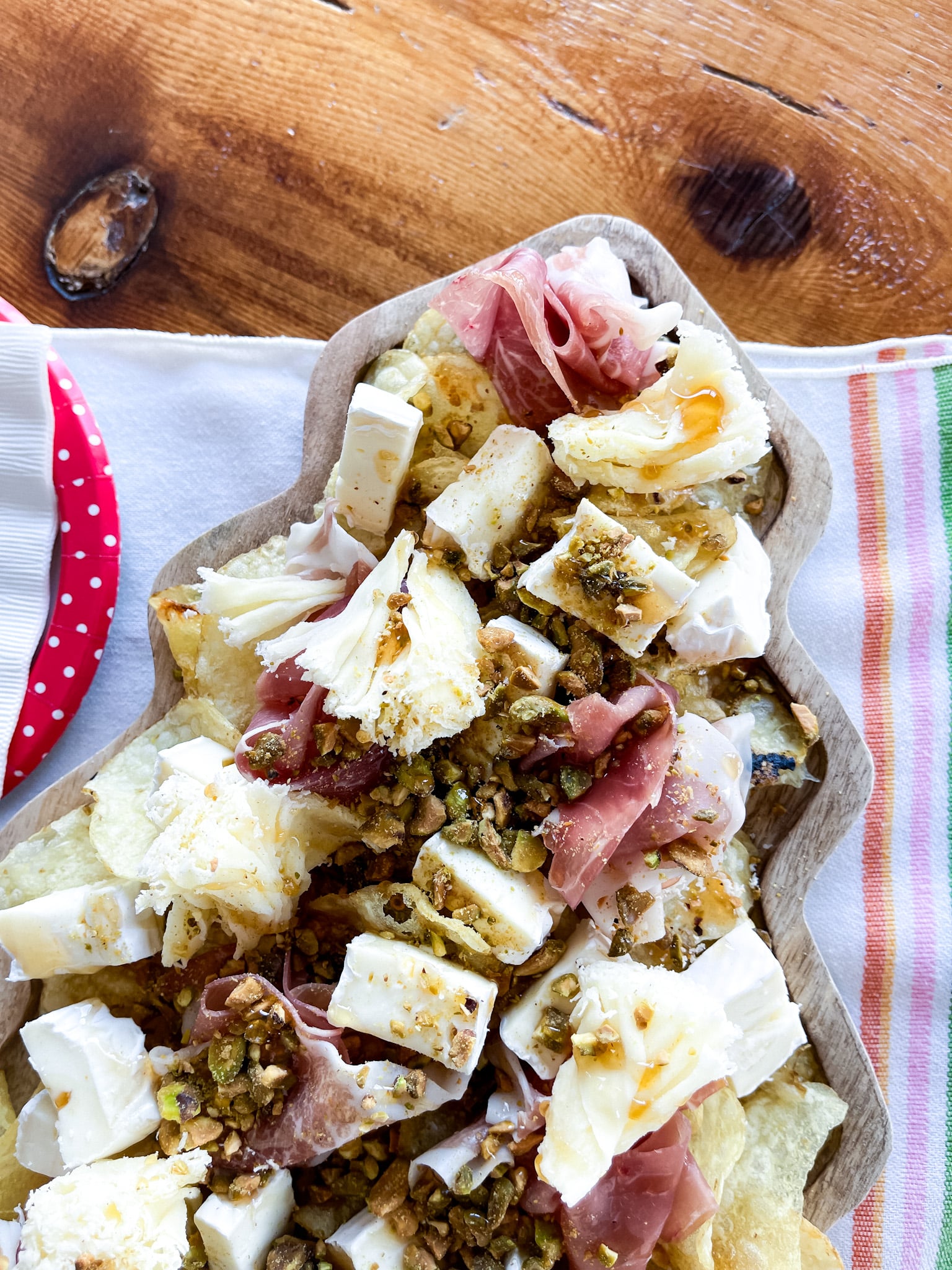 Tree shaped serving board with chipcuterie on it, sitting on a farmhouse table on a towel with vintage plates and napkins. Ingredients from Trader Joe's: potato chips, brie, prosciutto, pistachios, hot honey, tete de moine rosettes