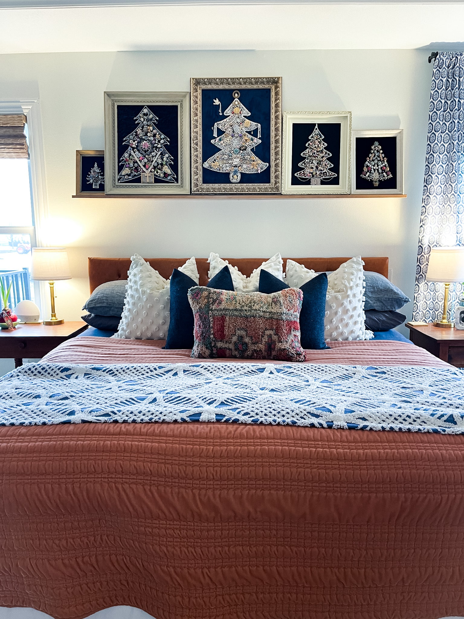 Bed with a faux leather headboard with lots of pillows and a picture ledge with vintage christmas jewelry tree art on it.