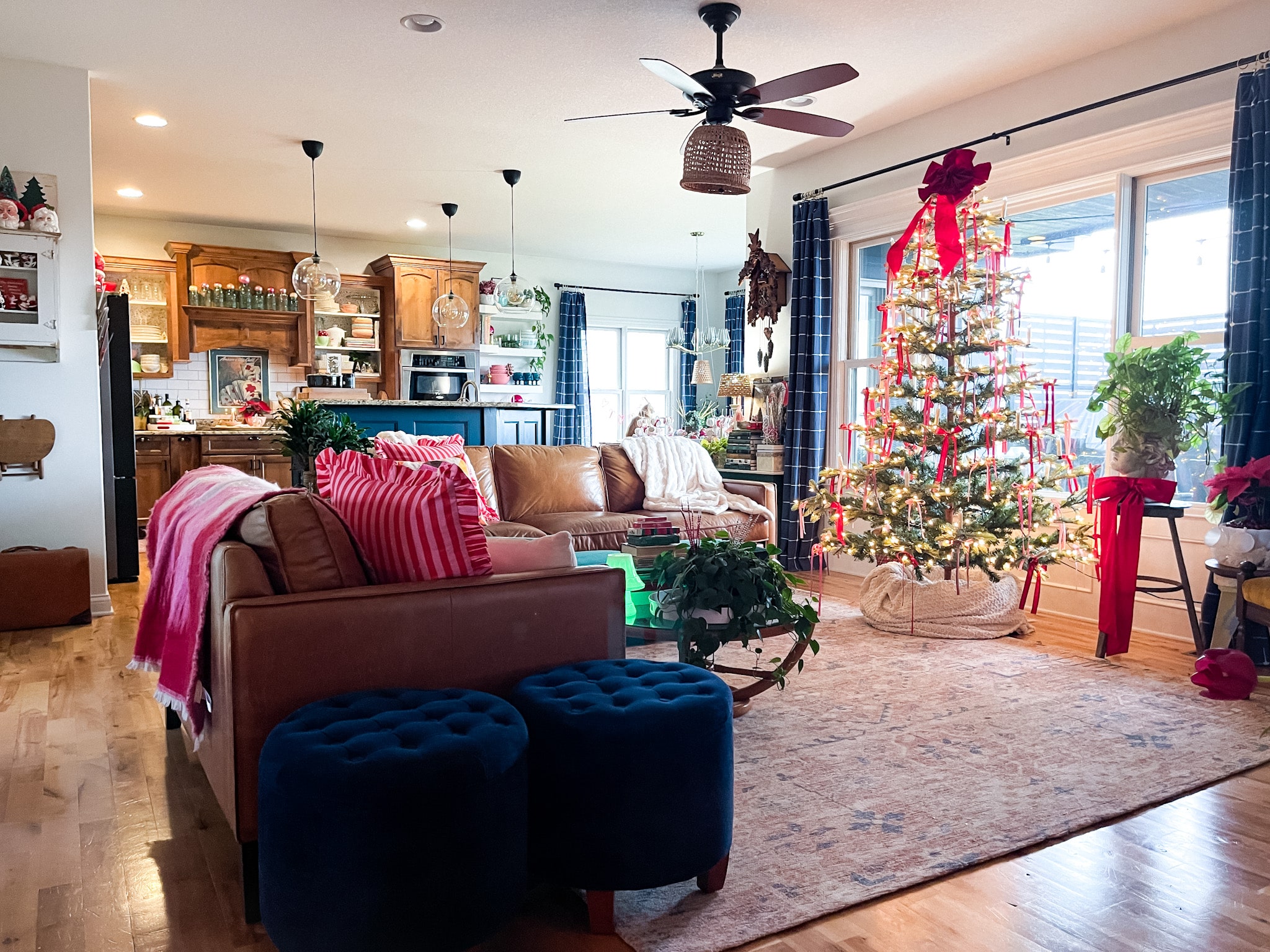 open concept living room and kitchen decorated for christmas. a couple of leather sofas, a christmas tree decorated with pink and red ribbons, and two blue footstools