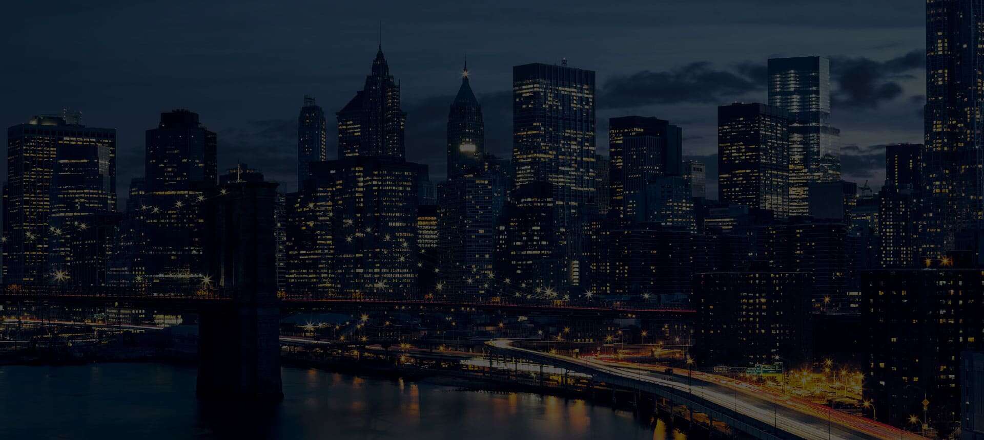 City skyline at night with illuminated skyscrapers and a lighted bridge over a river.