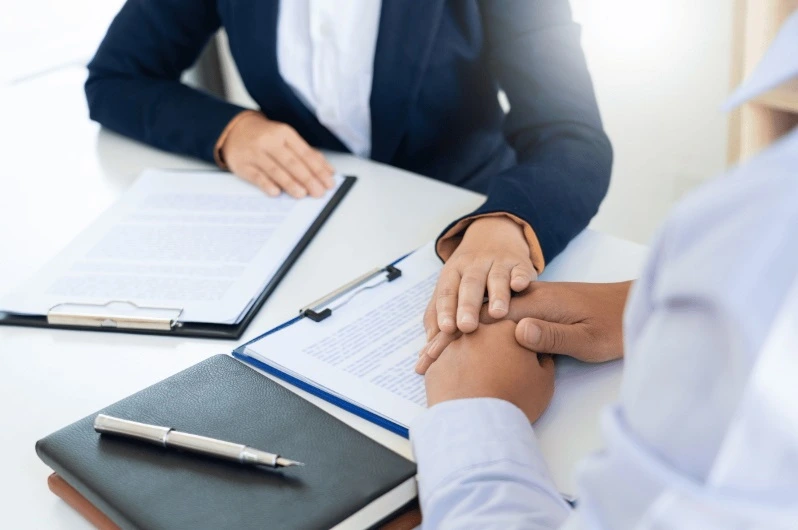 Close-up of a person in a navy suit placing their hand in a gesture of support and comfort over the hand of another person, with legal documents and a pen on a table between them.