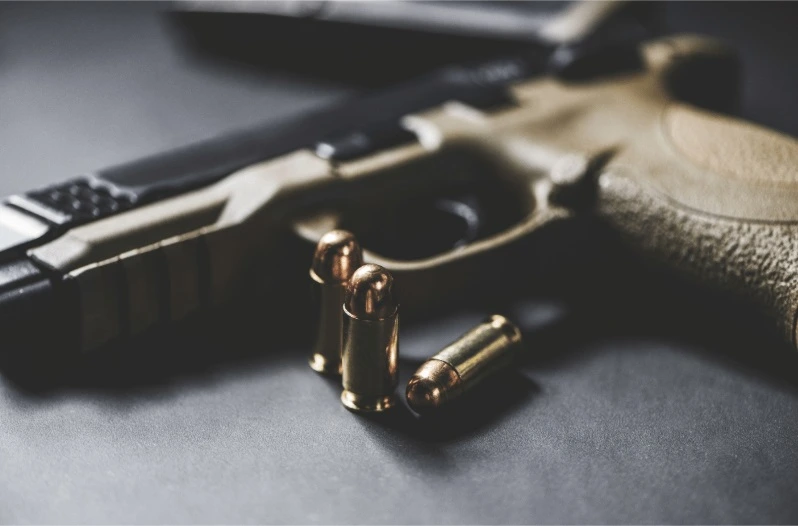 A close-up, low-light view of a semi-automatic handgun and three copper-jacketed bullets (ammunition) on a dark surface.